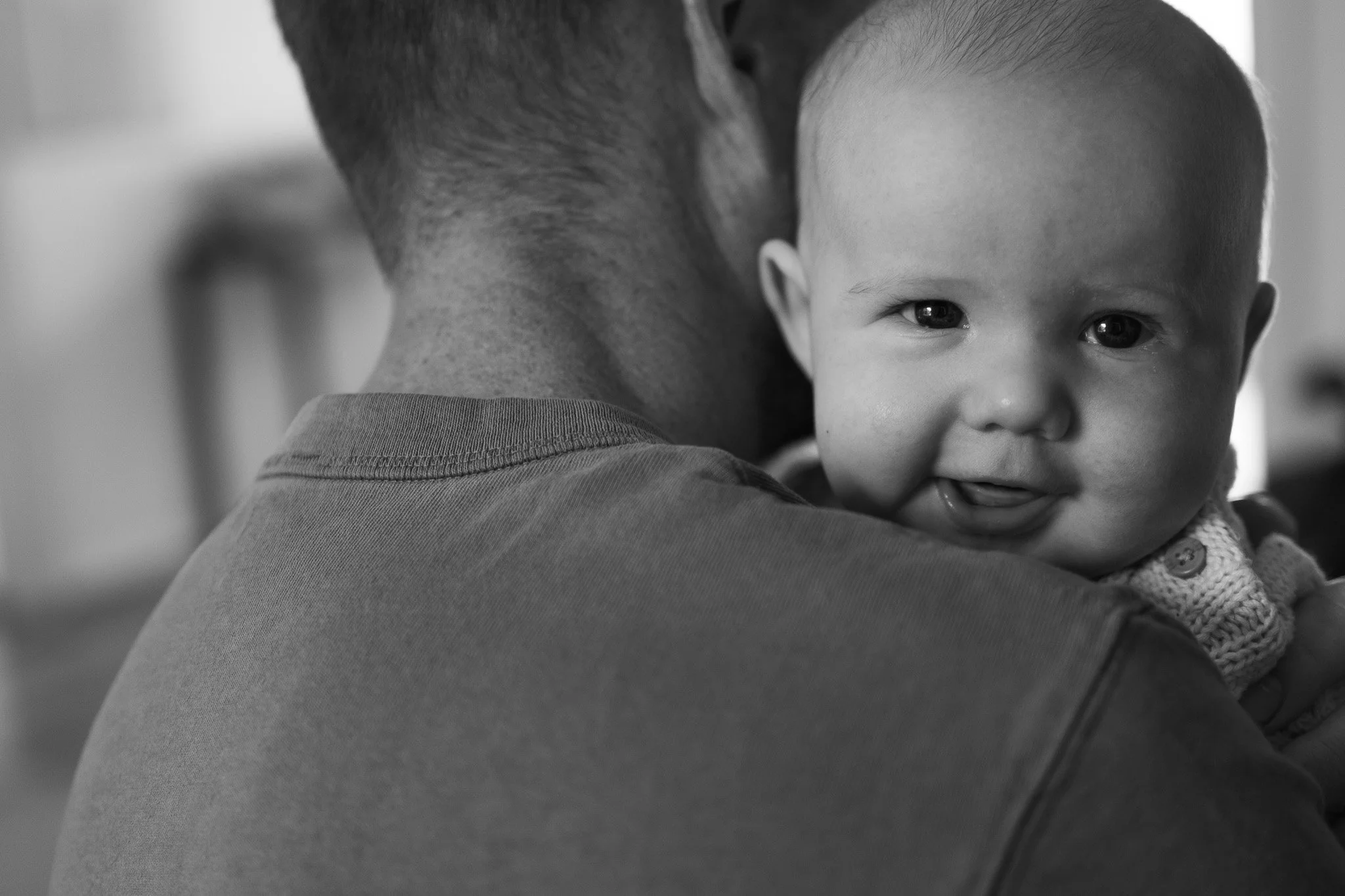 A child being held closely in front of an adult, with the child smiling and looking directly at the camera. The photo is in black and white.