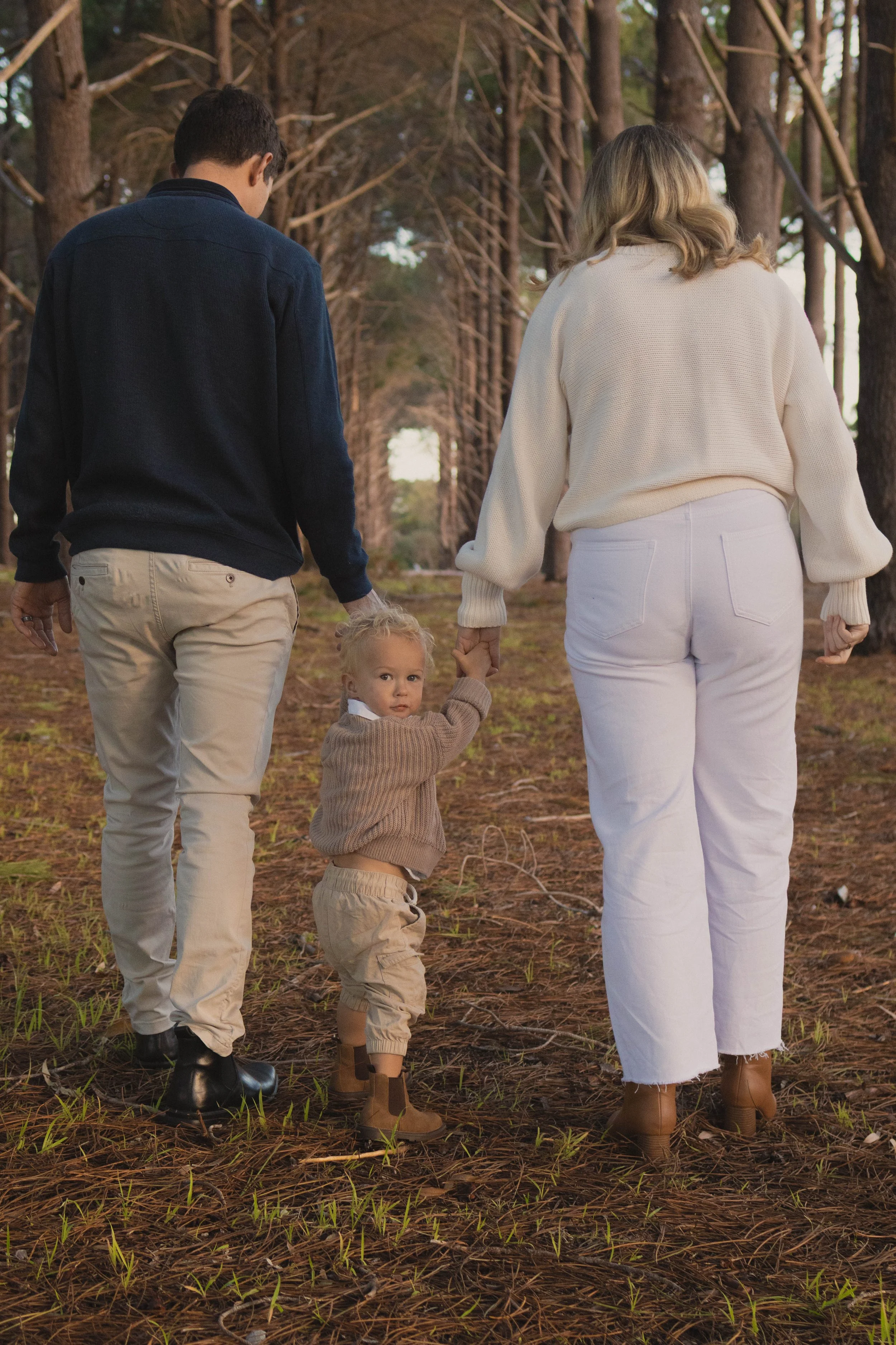 A family of three walking through a forest with tall trees, holding hands. The child is looking up with curiosity, wearing beige pants, a brown sweater, and brown boots. The parents are dressed casually, with the father in beige pants and a dark jack