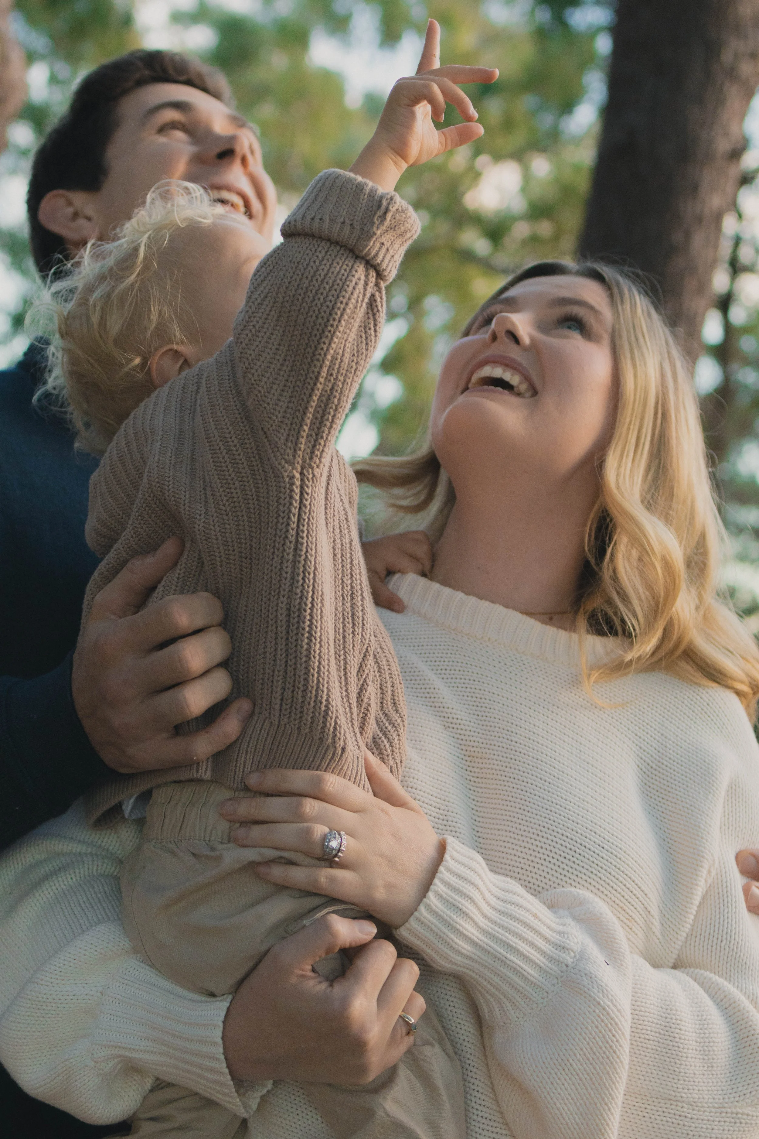 A family of four enjoying outdoor time, with the young girl reaching up and the mother looking up happily, surrounded by trees.