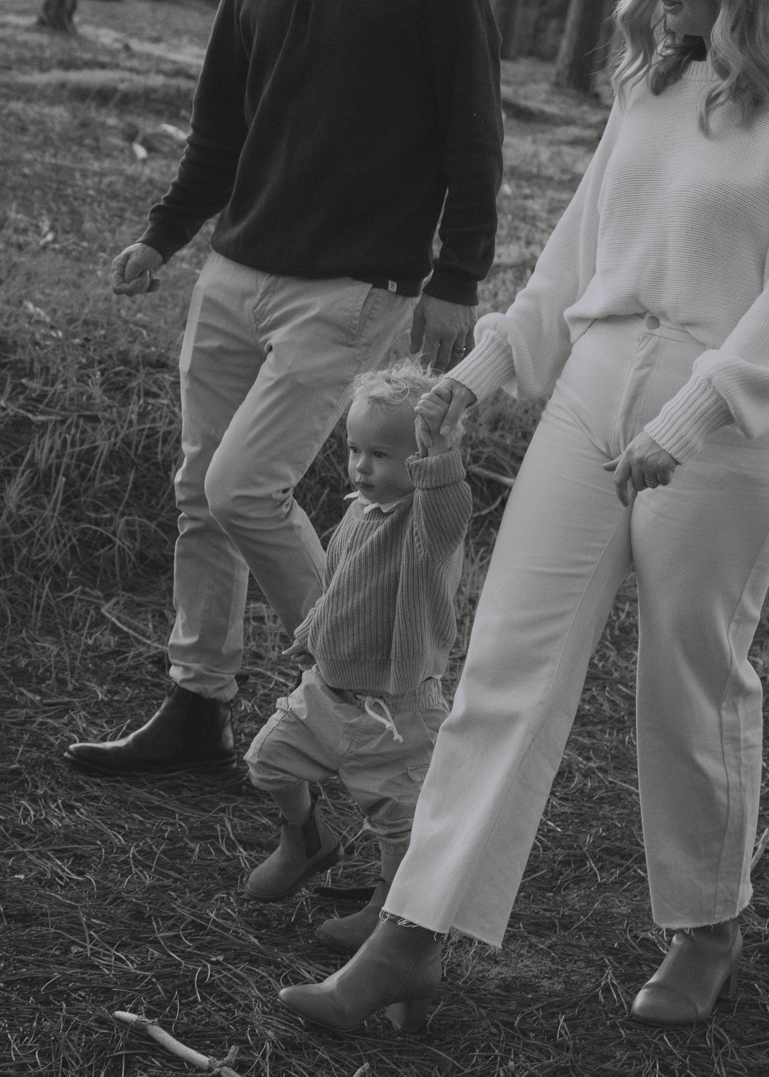 A black and white photo of a child holding hands with two adults, walking outdoors in a field with grass and trees.