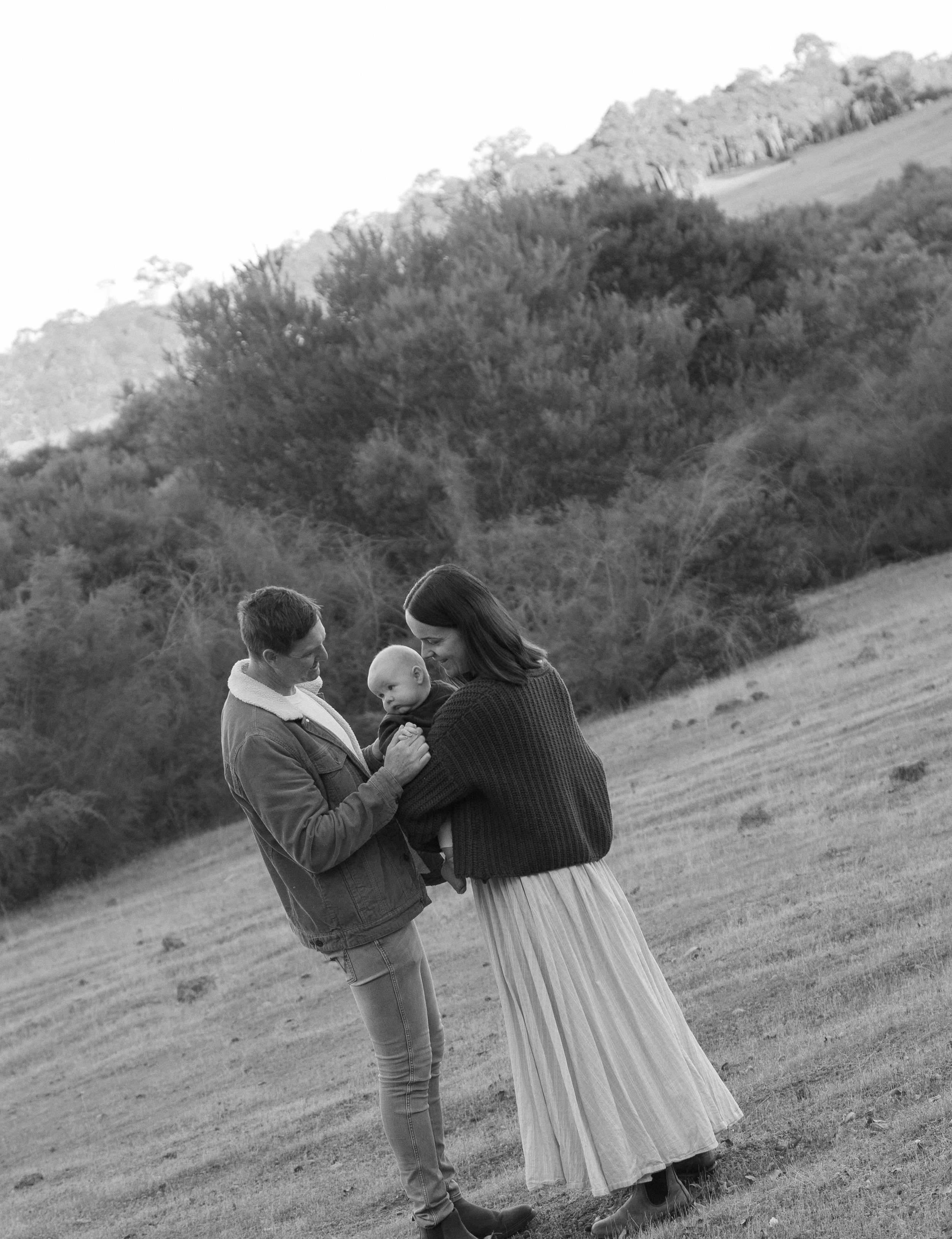 A man and woman are standing outdoors, holding hands, with a baby between them. The man is wearing a jacket and jeans, the woman is wearing a long skirt and a sweater, and the baby is looking at the woman. There are trees and rolling hills in the bac