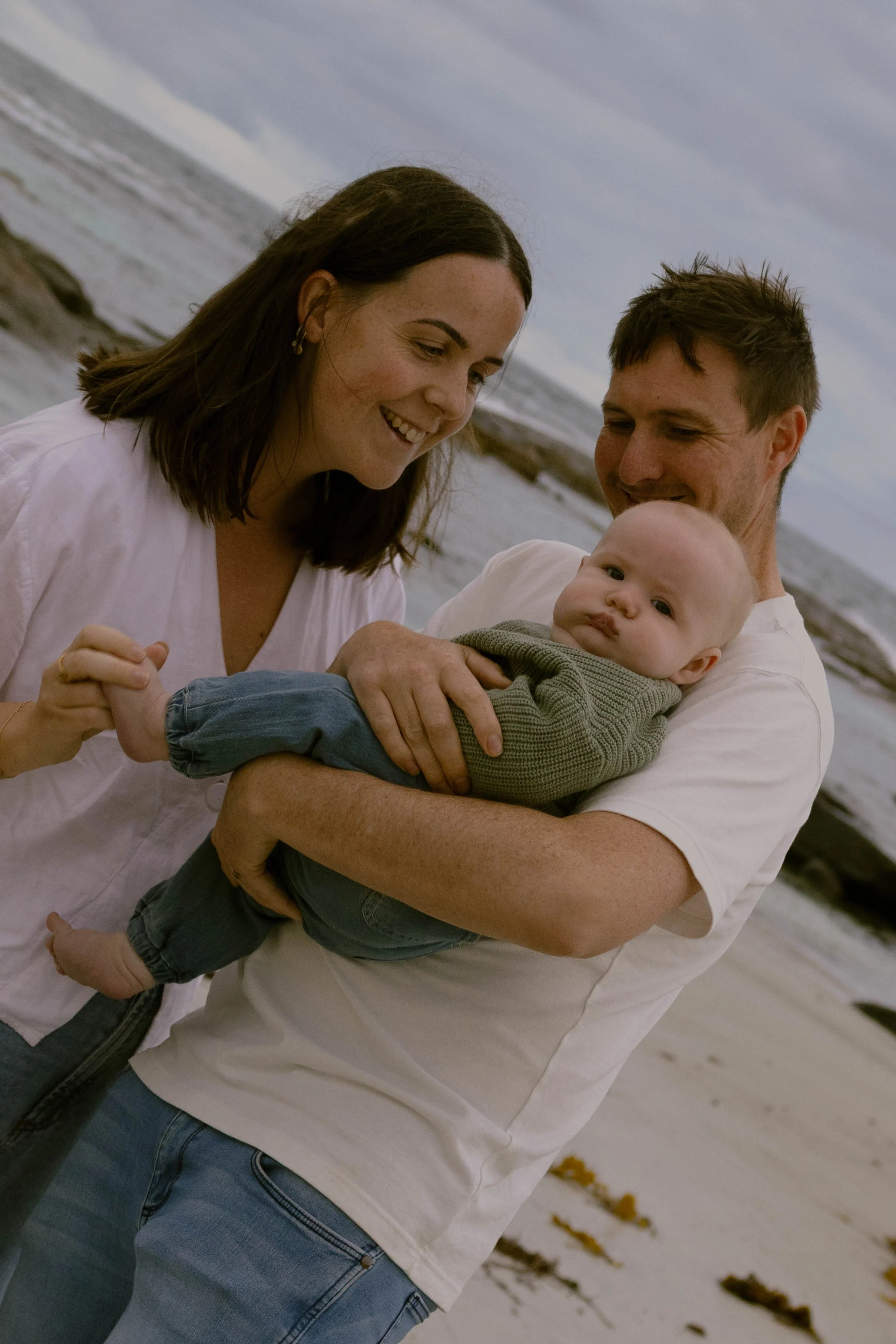 A man and woman holding a baby on a beach during cloudy weather.