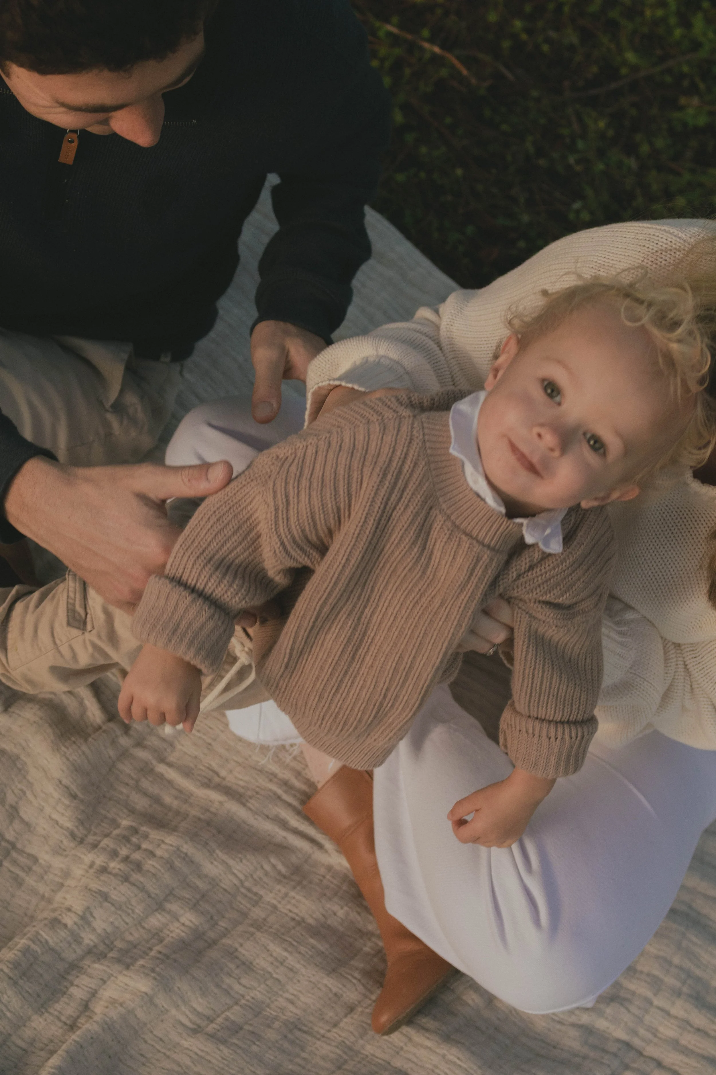 A young boy with curly blonde hair, wearing a beige sweater over a collared shirt, is lying on a bed with a white sheet and a brown leather ankle boot. An adult in a black sweater is holding the boy's arm. The scene is indoors with a textured carpete