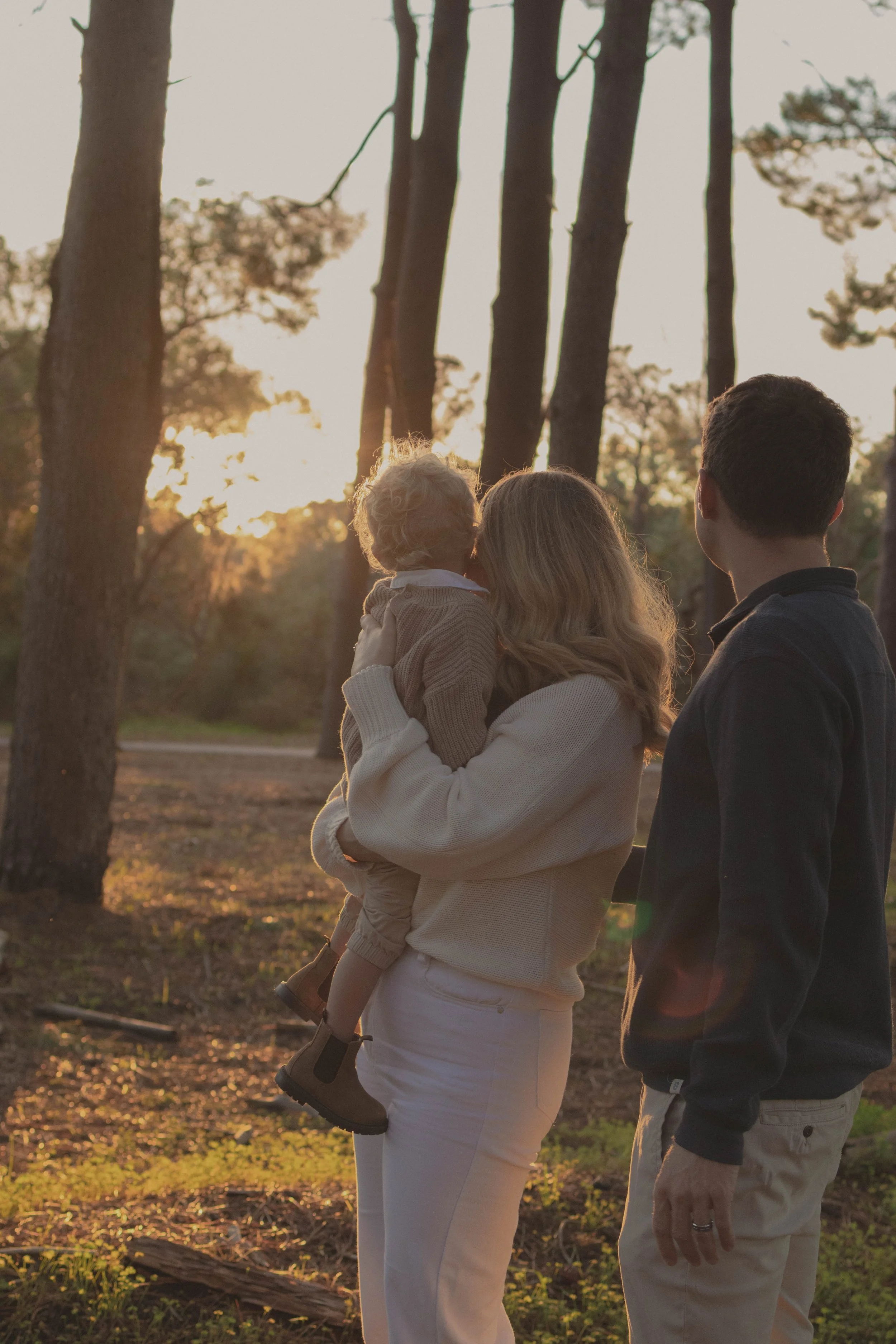 A woman holding a young child in her arms while standing outdoors during sunset, with a man standing beside her, all looking at the trees and sunset in the background.
