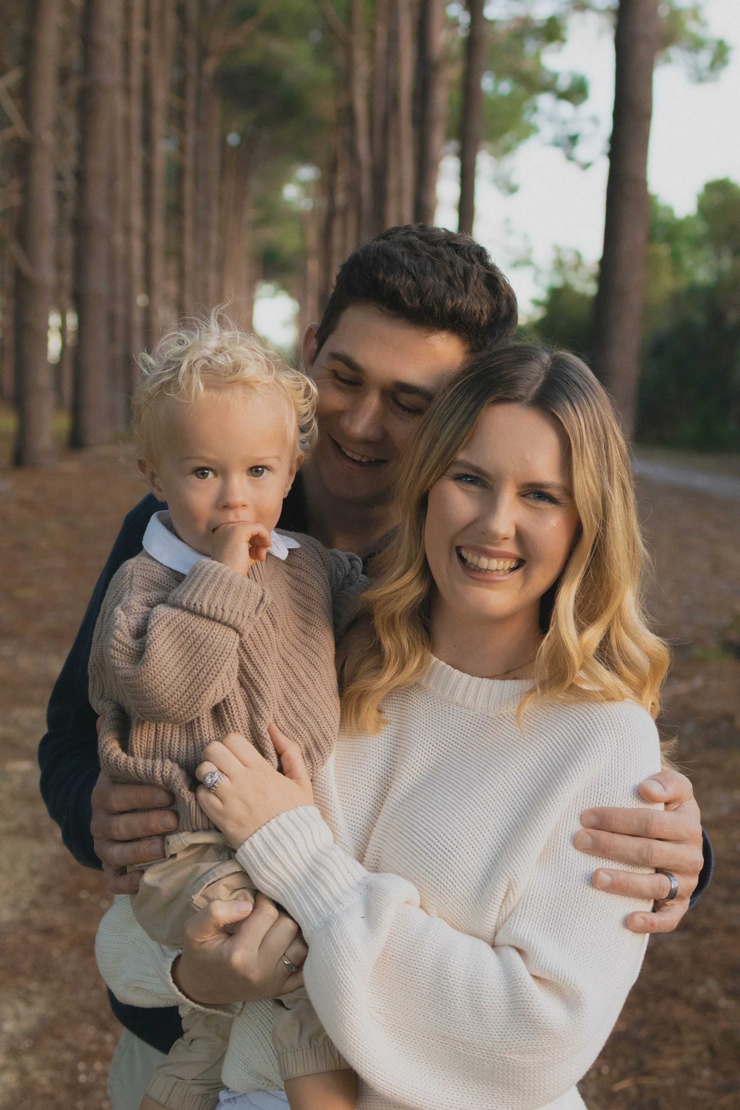 A happy family of three, including a woman, a man, and a young boy, posing together outdoors in a wooded area during daytime. The woman has long blonde hair and is smiling, while the man and boy are close together, with the boy sucking his thumb.