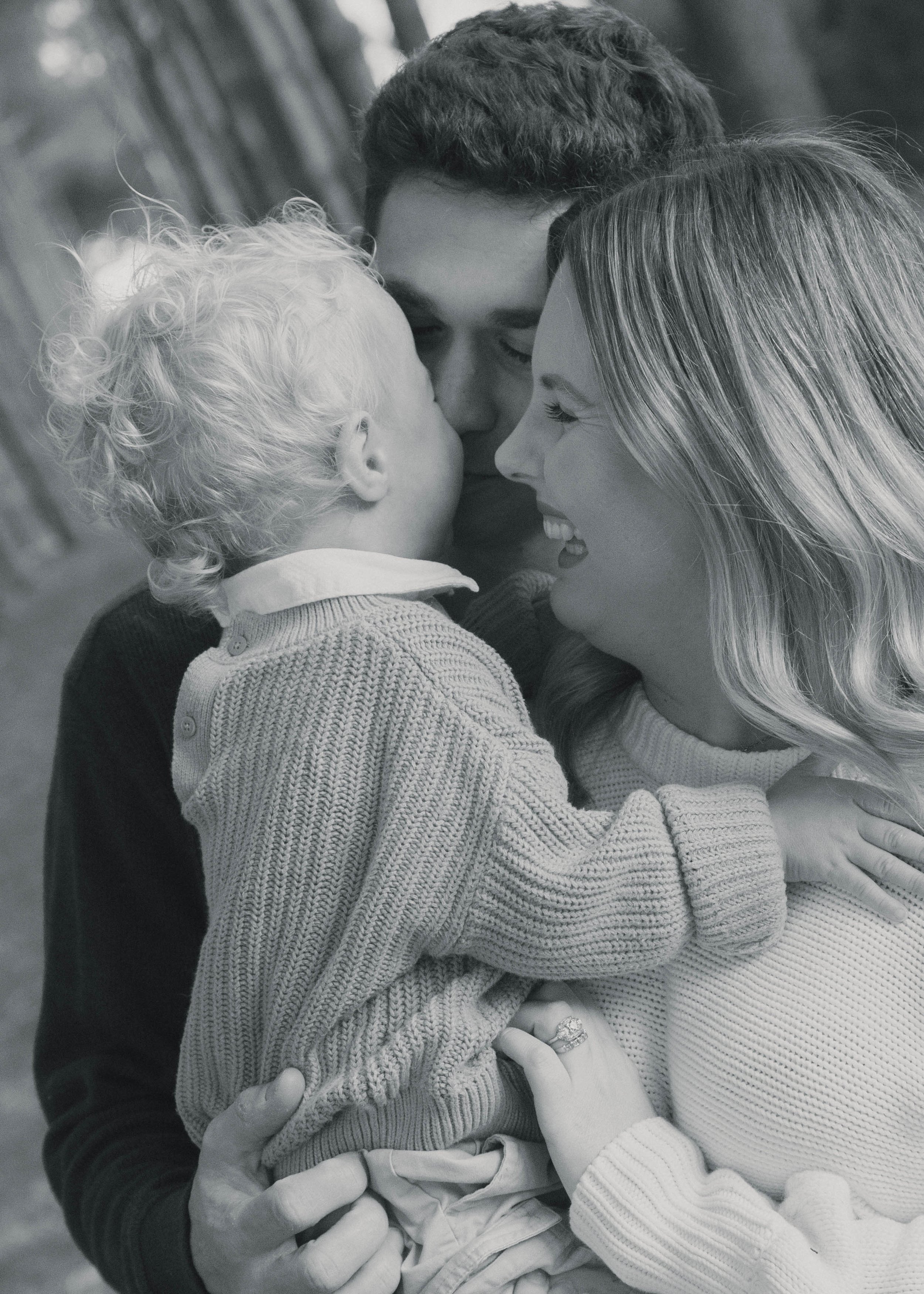 A family of three, including a young child, a woman, and a man, sharing a close, affectionate moment outdoors. The child and woman are touching noses, and they are all smiling.