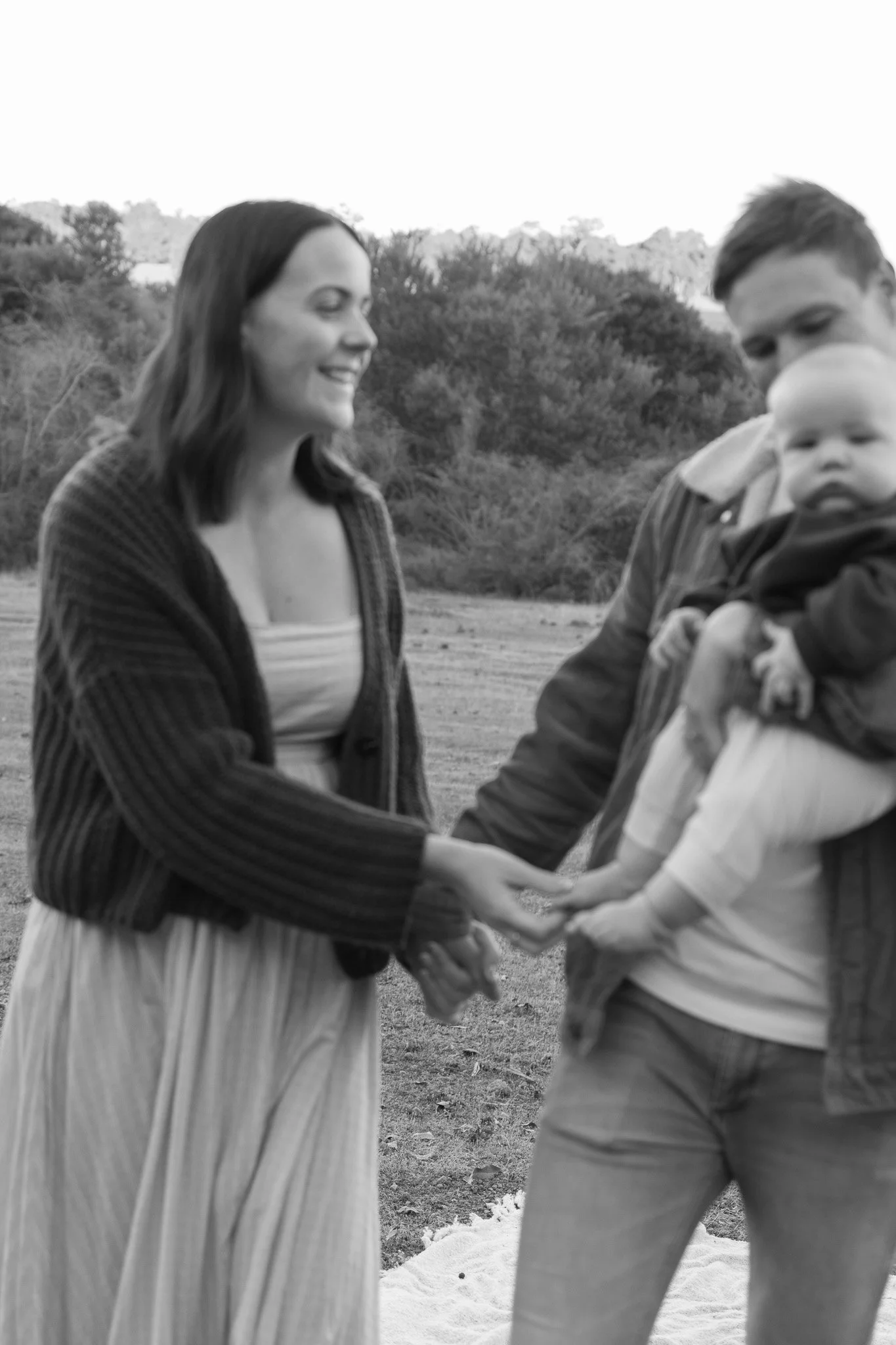 A woman and a man holding a baby outdoors, smiling and looking at each other, with trees in the background.