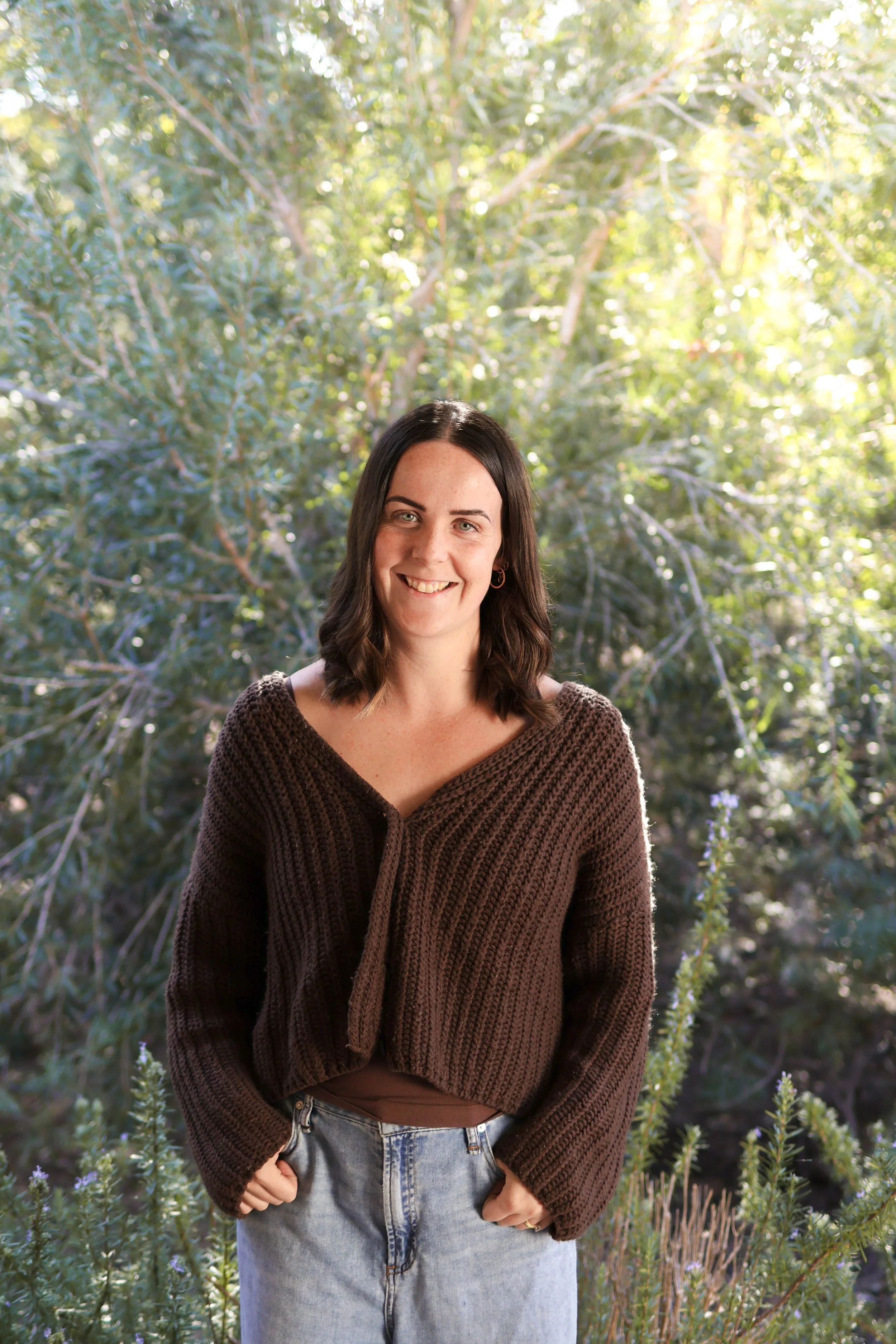 A woman with dark hair, smiling, wearing a brown knitted cardigan and blue jeans, standing outdoors in front of greenery and trees.