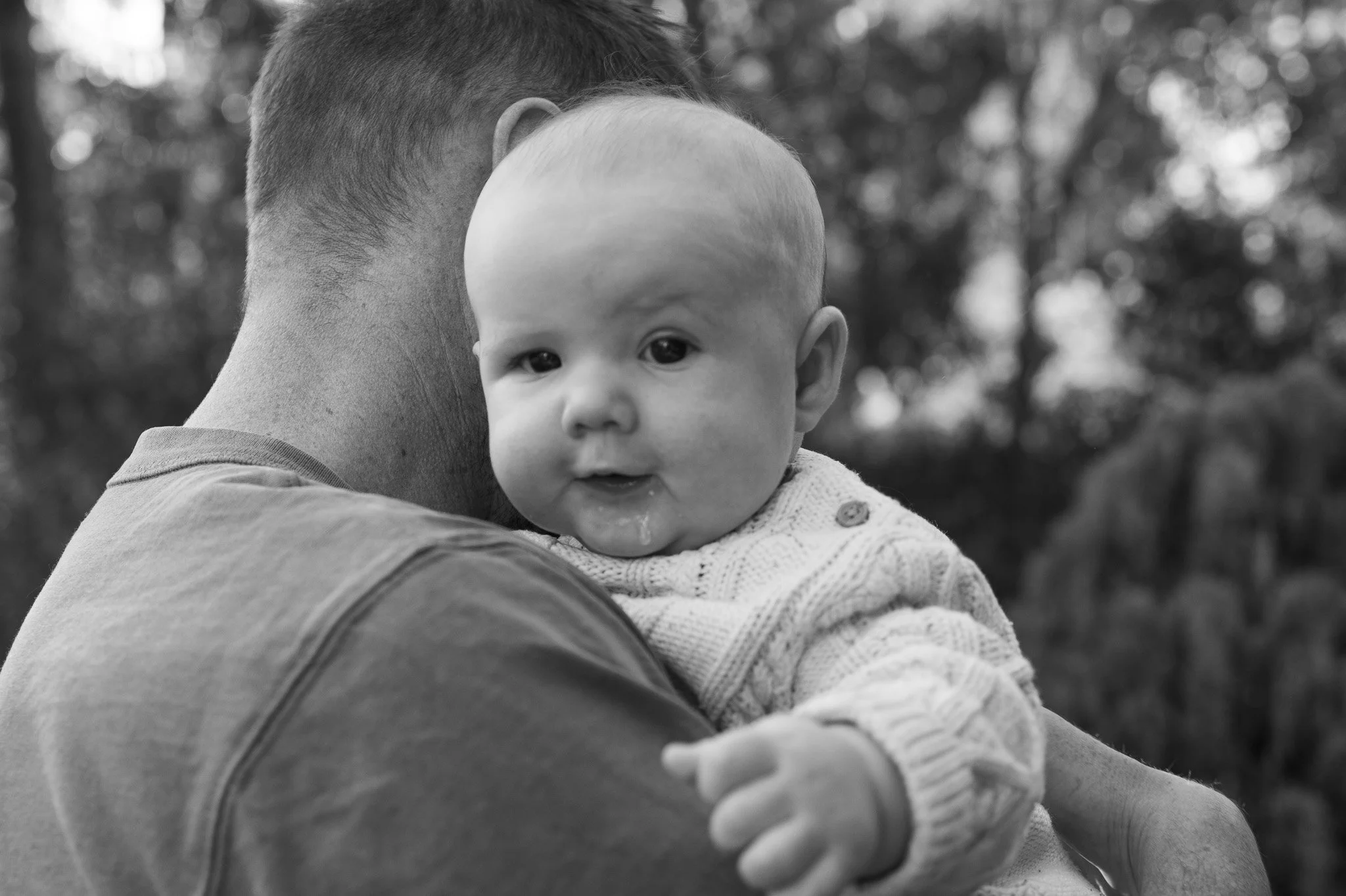 A person holding a baby outdoors, with trees blurred in the background. The photo is in black and white.