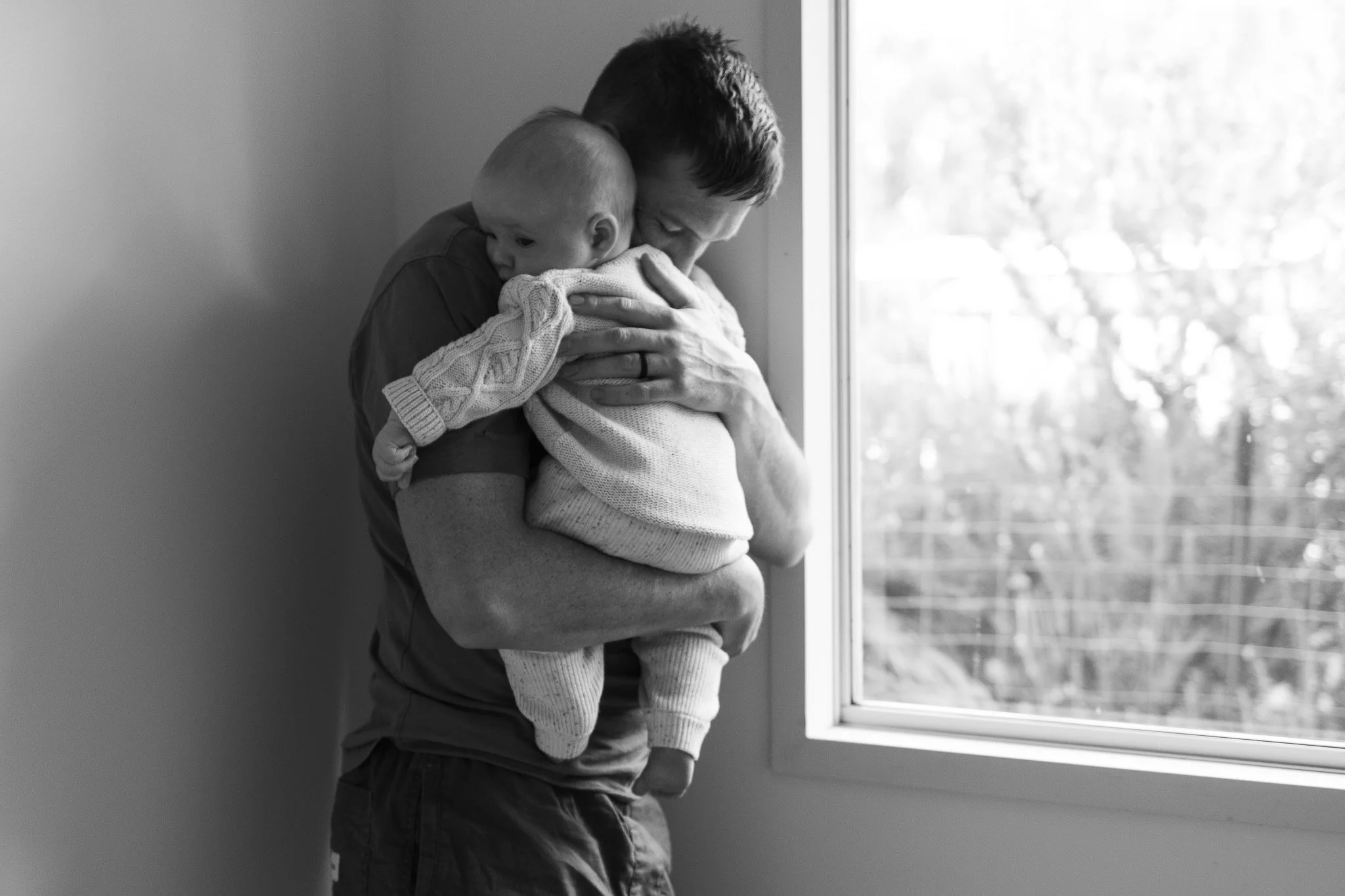 A man holding a young child while standing by a window, with the child resting their head on the man's shoulder; the man appears to be embracing or comforting the child.