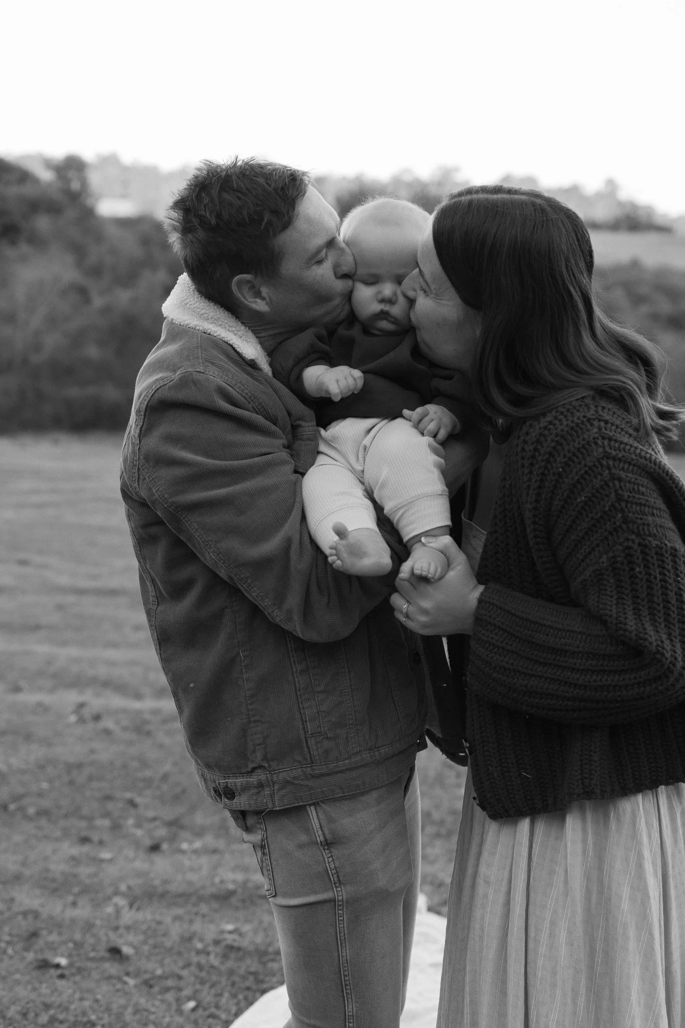 A black and white photo of a man and woman holding a baby, kissing the child's cheeks in an outdoor setting.