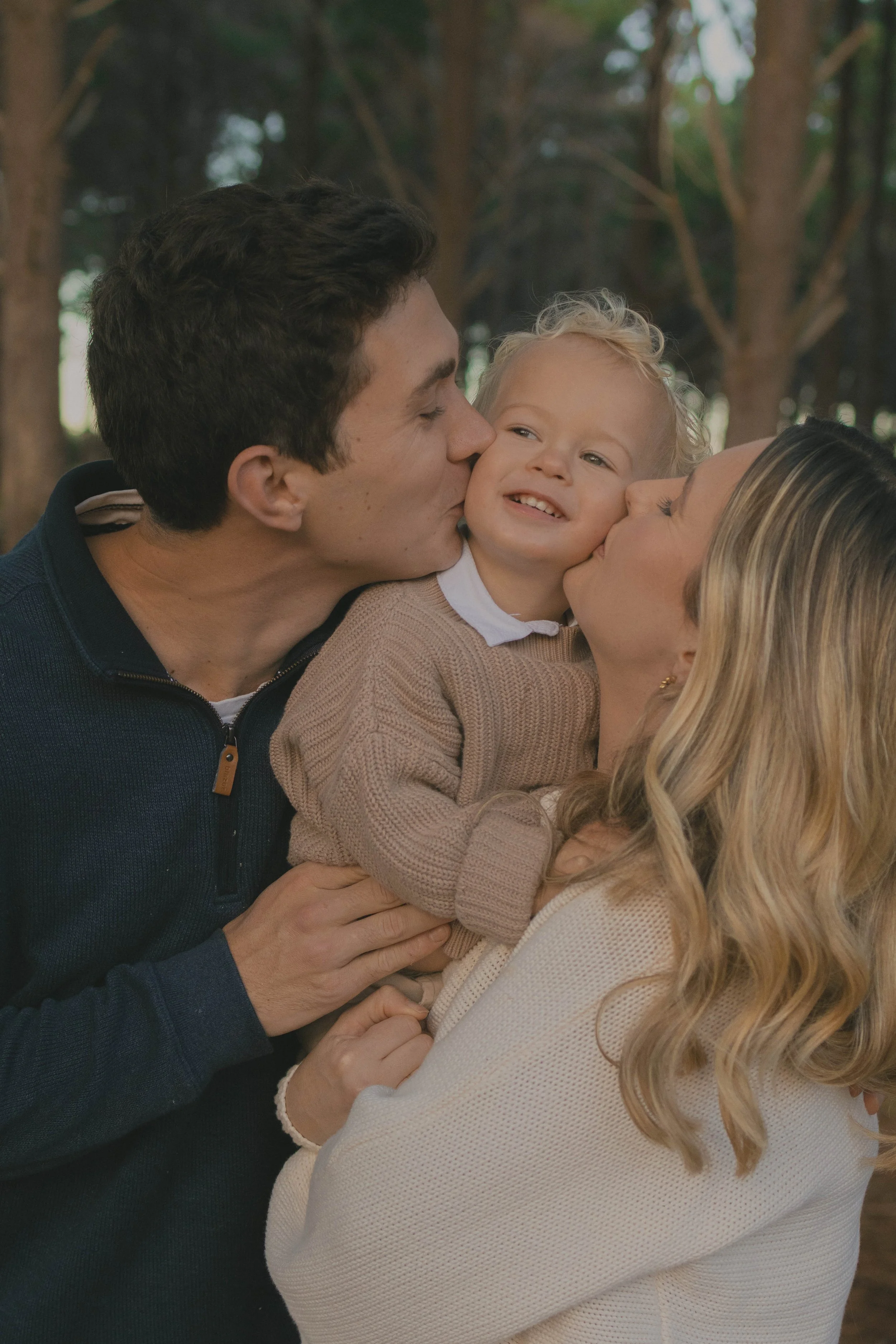 A happy family of three sharing a moment outdoors, with the father kissing the child on the cheek and the mother holding the child, all in a wooded area.
