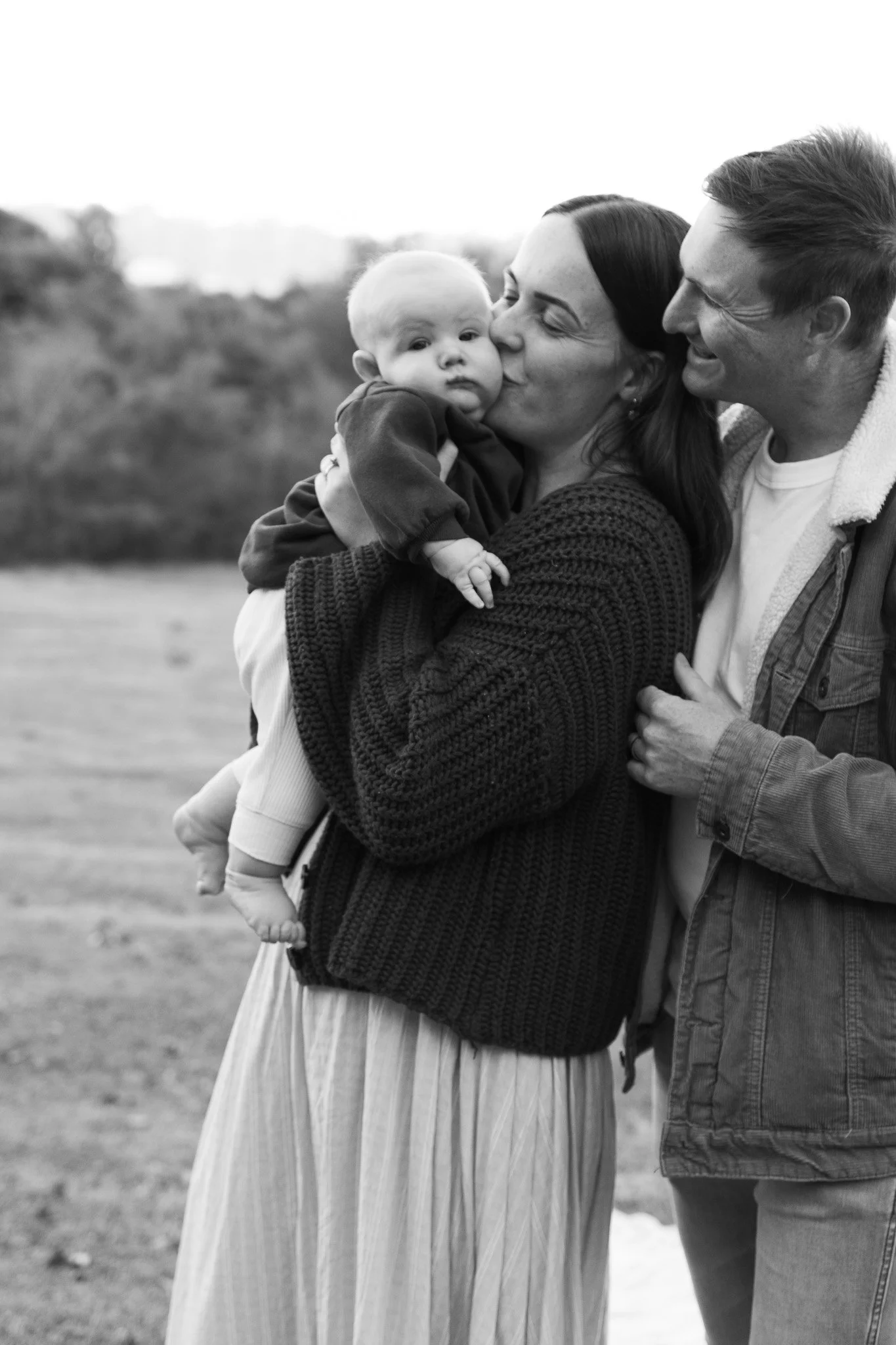 A black and white photo of a happy family outdoors. The mother is kissing her baby, who is being held by another person, possibly the father. The scene shows affection and joy.