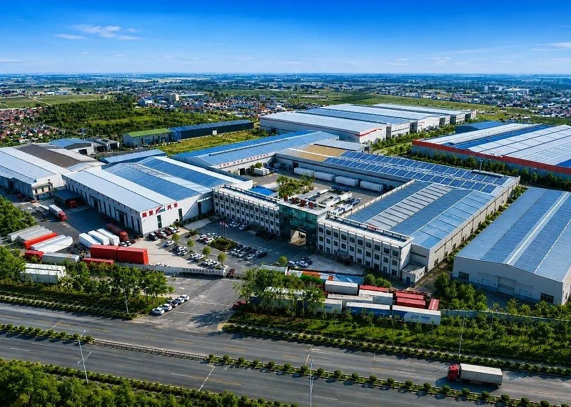 Aerial view of a large industrial complex with warehouses, loading docks, trucks, and parking lots, situated along a highway with trees and fields in the background.