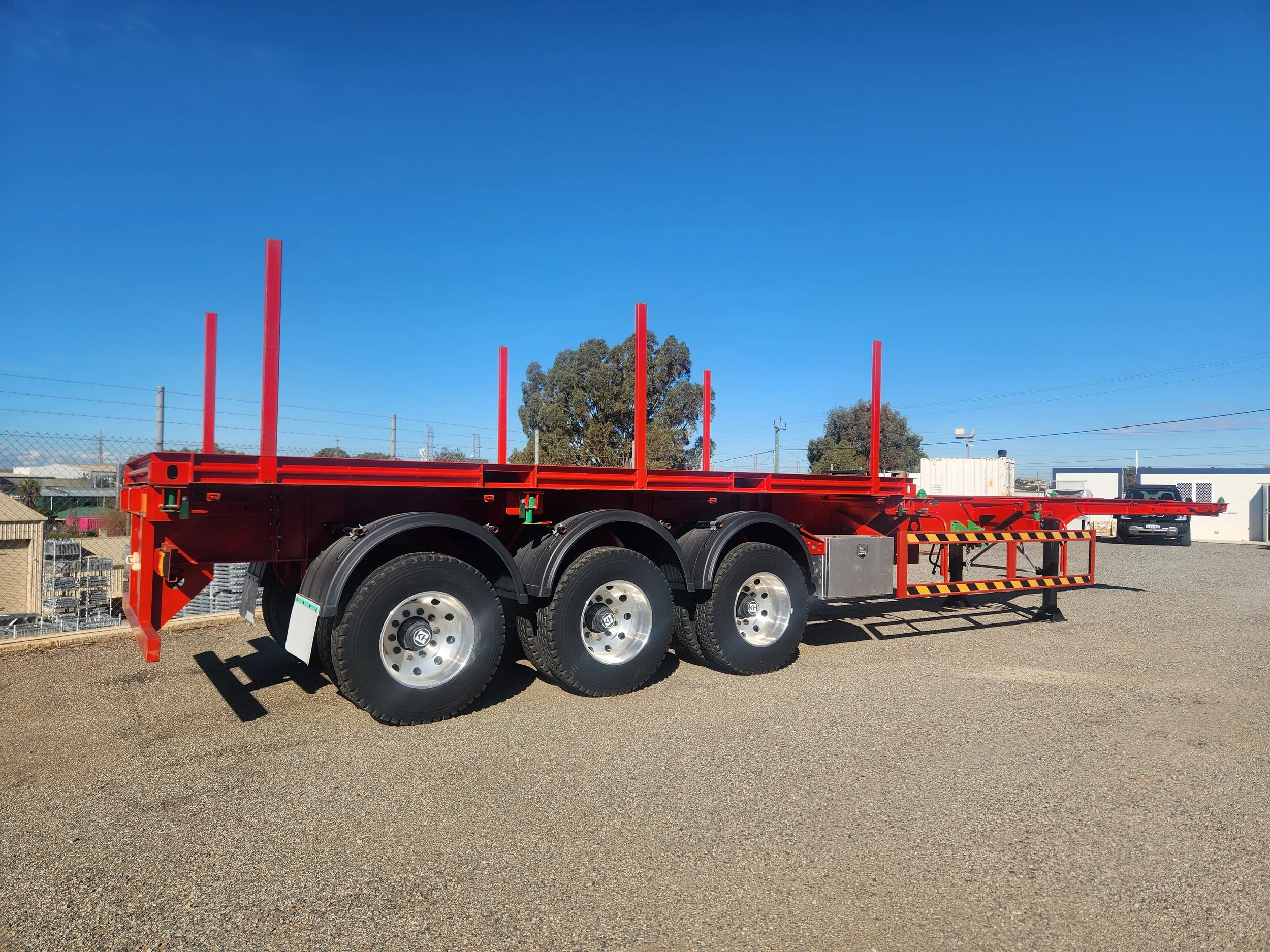 Red flatbed truck trailer parked outdoors under a clear blue sky with trees, fences, and other industrial elements in the background.