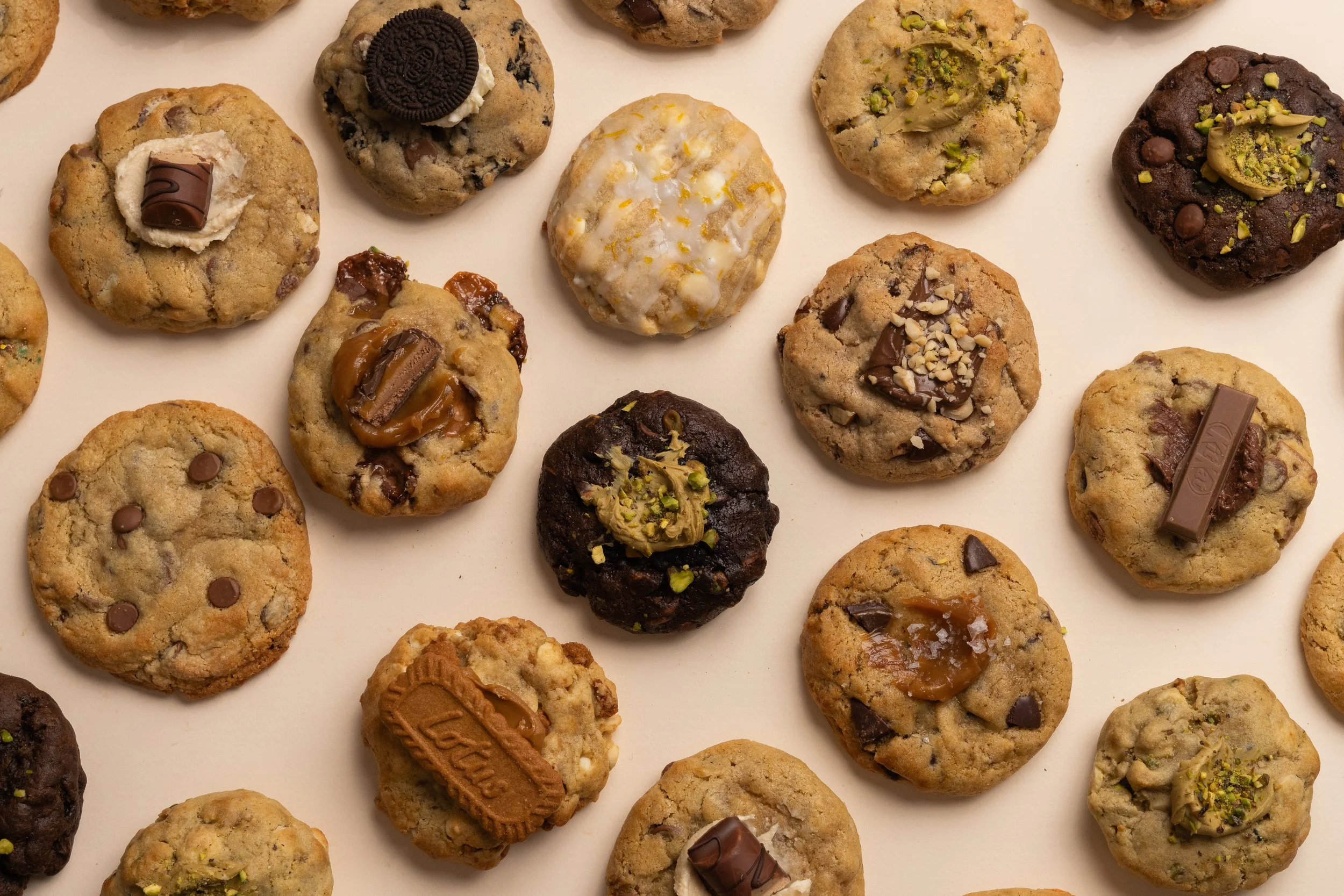A variety of chocolate chip cookies, some topped with nuts, pistachios, or chocolate pieces, arranged on a white surface.