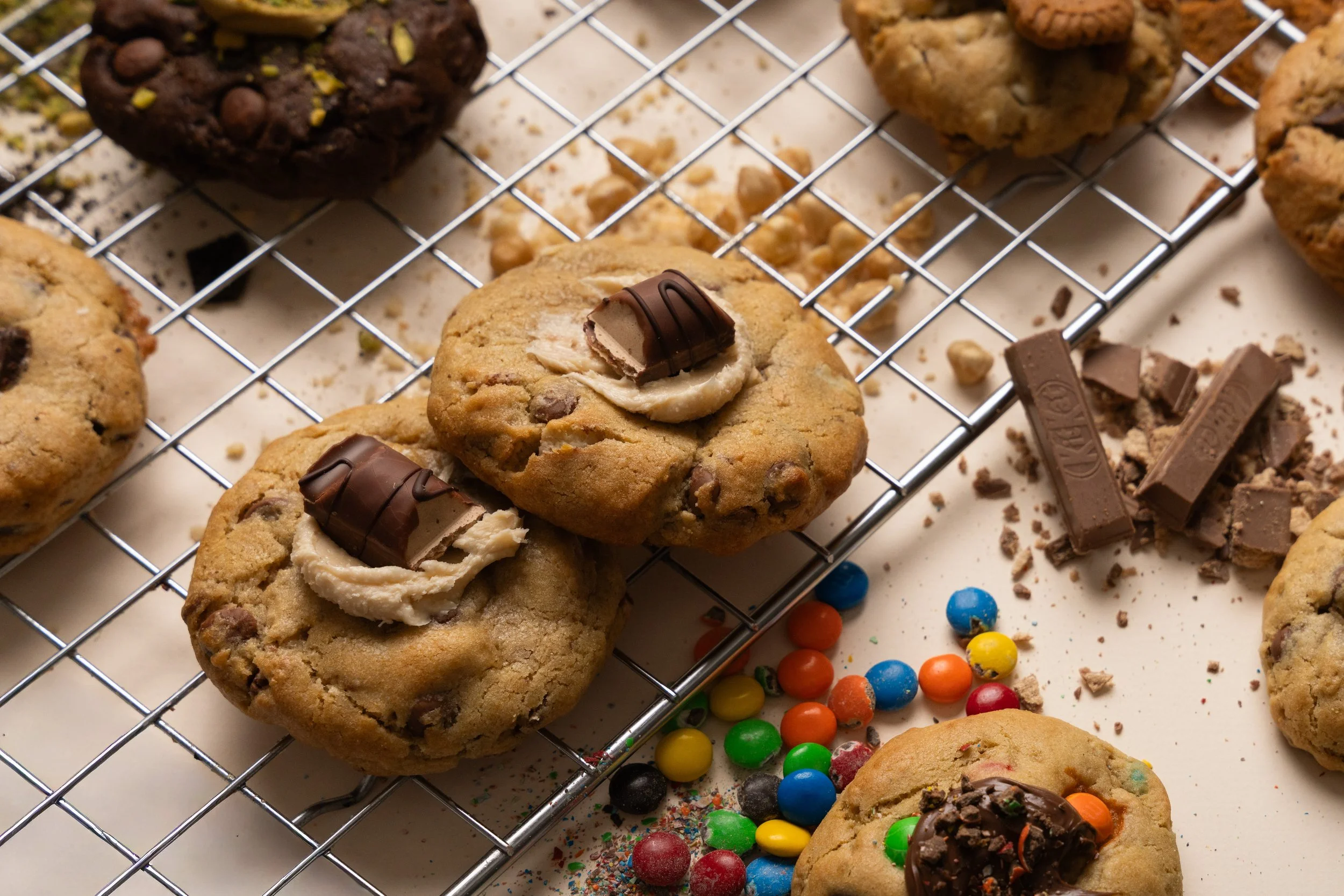 Assorted cookies on a cooling rack with colorful candy accents around. The cookies include chocolate chip, peanut butter with chocolate on top, and oatmeal raisin with nuts. Some cookie crumbs and broken pieces of chocolate are scattered on the surface below.