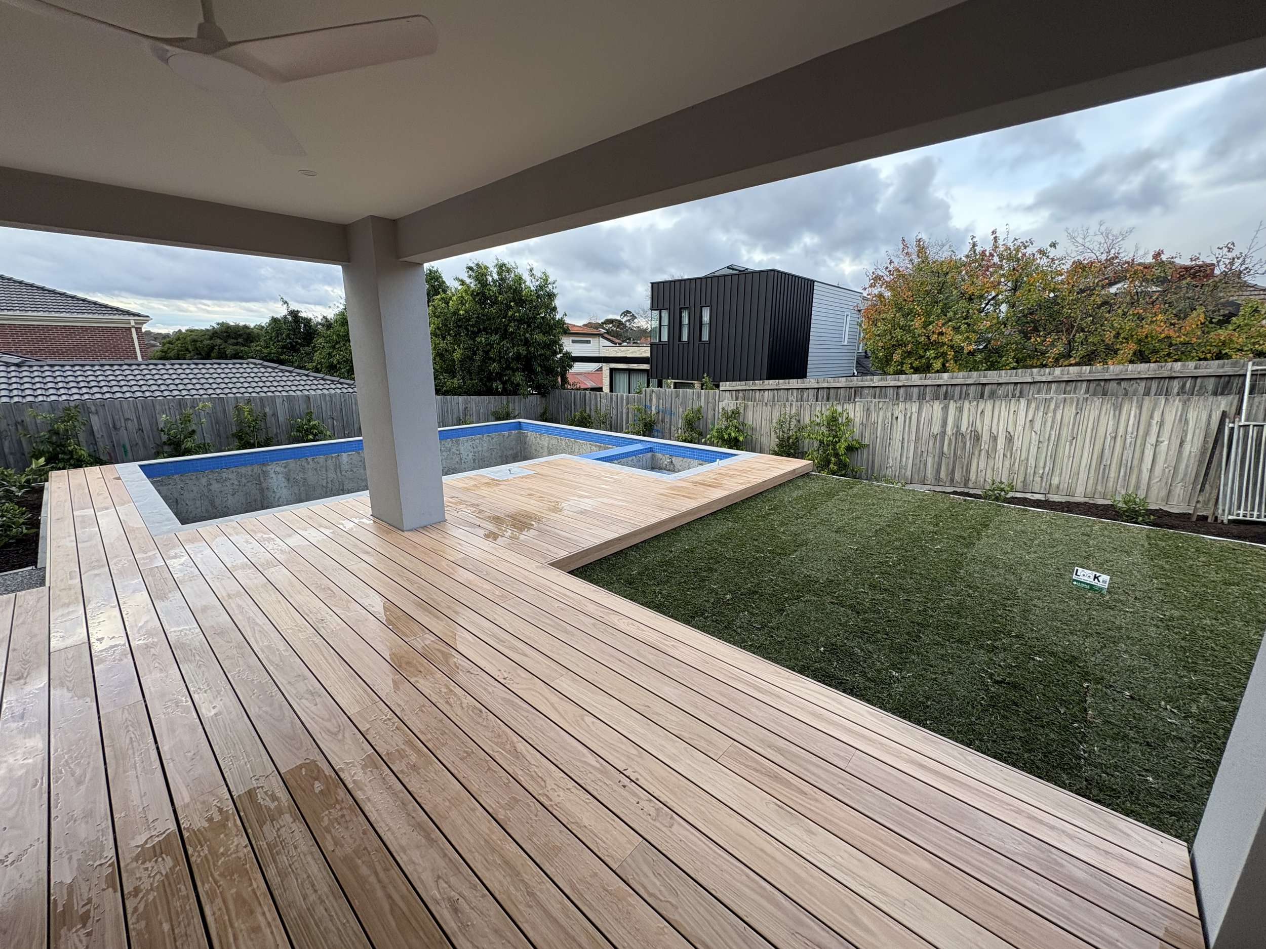 A backyard patio with a small in-ground pool, wooden deck, and a grassy area with a fence and trees in the background. The sky is cloudy.