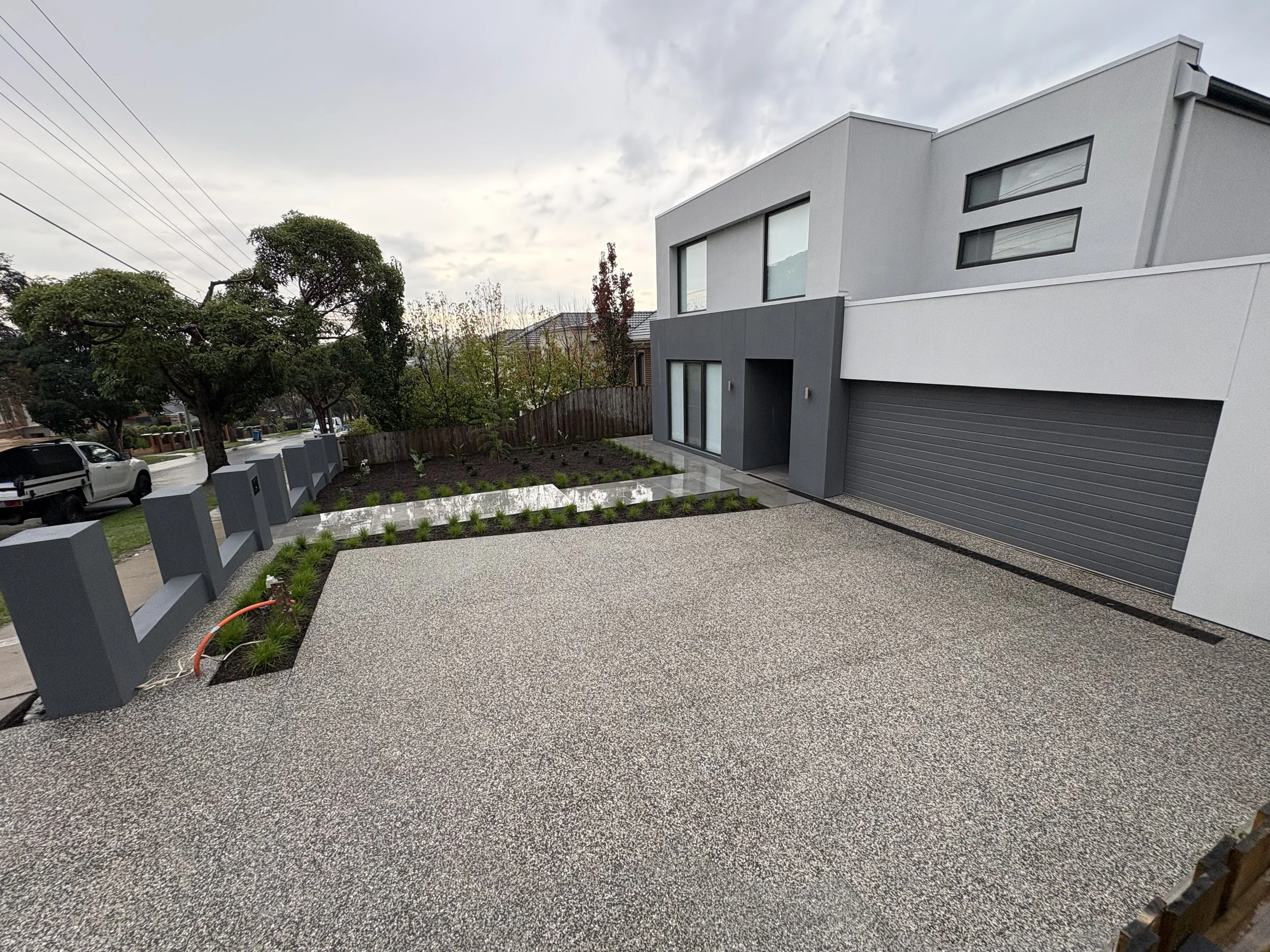 Modern white and gray two-story house with a minimalist design, large windows, a garage, and a small landscaped yard with plants and trees, under a cloudy sky.