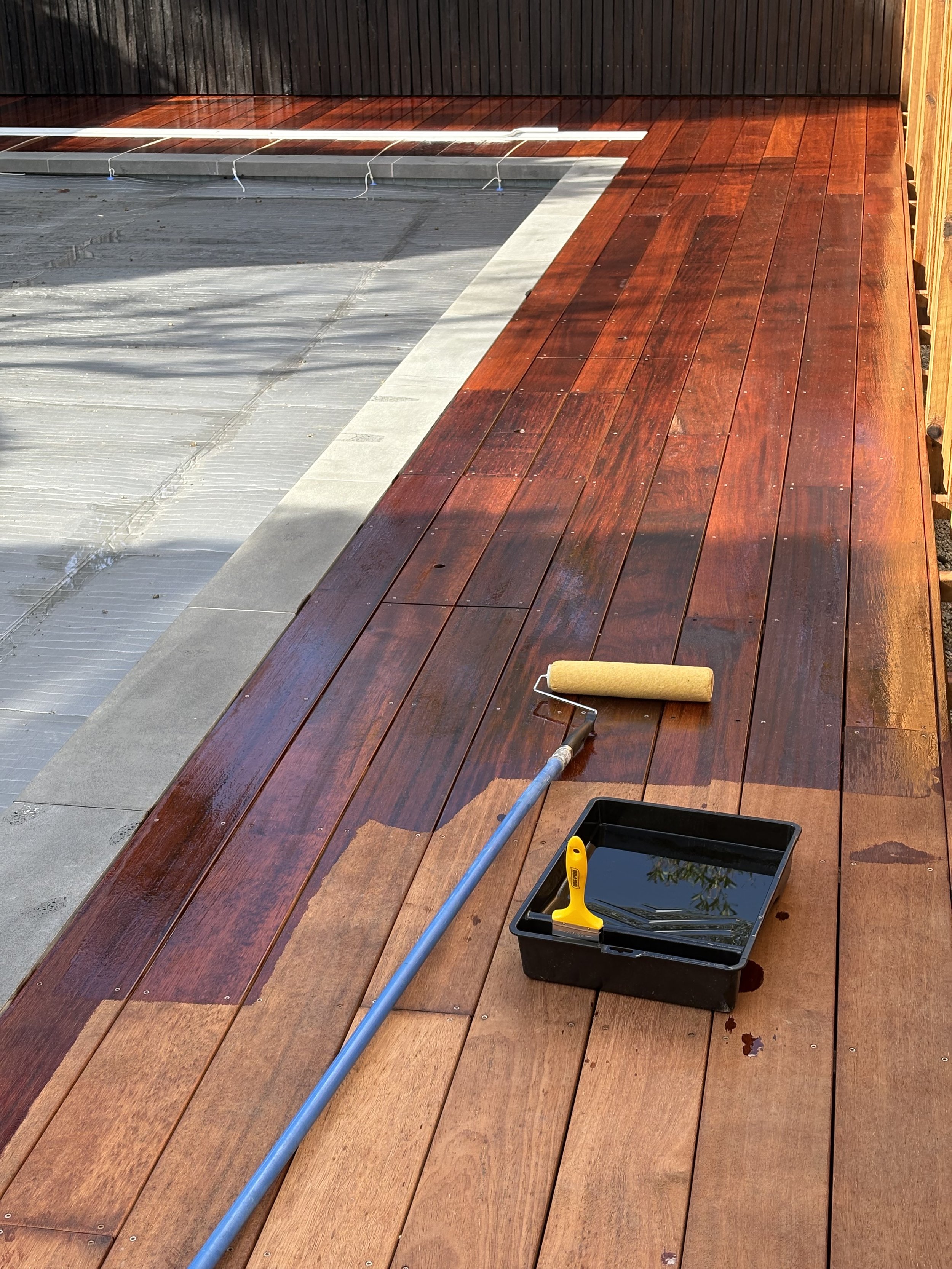 Wooden deck being stained with a paint roller, a paint tray with black stain, and a paint scraper on the tray. The deck is partially finished with a darker, stained section and unfinished lighter section.