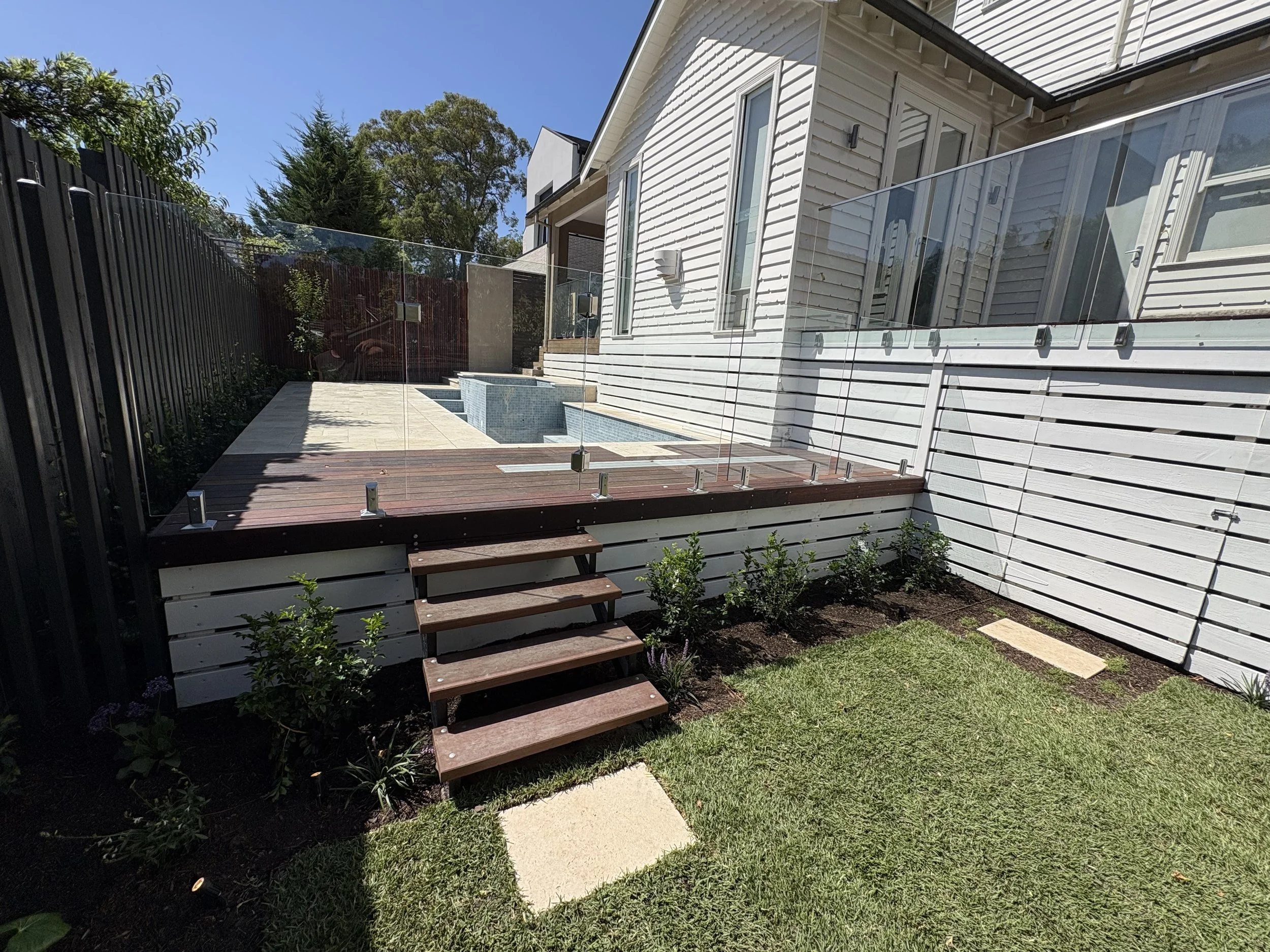 A backyard patio area with wooden steps leading down to a grassy yard, a small swimming pool with blue tiles, white wooden fence, and the side of a white house with large windows and glass balcony railing.