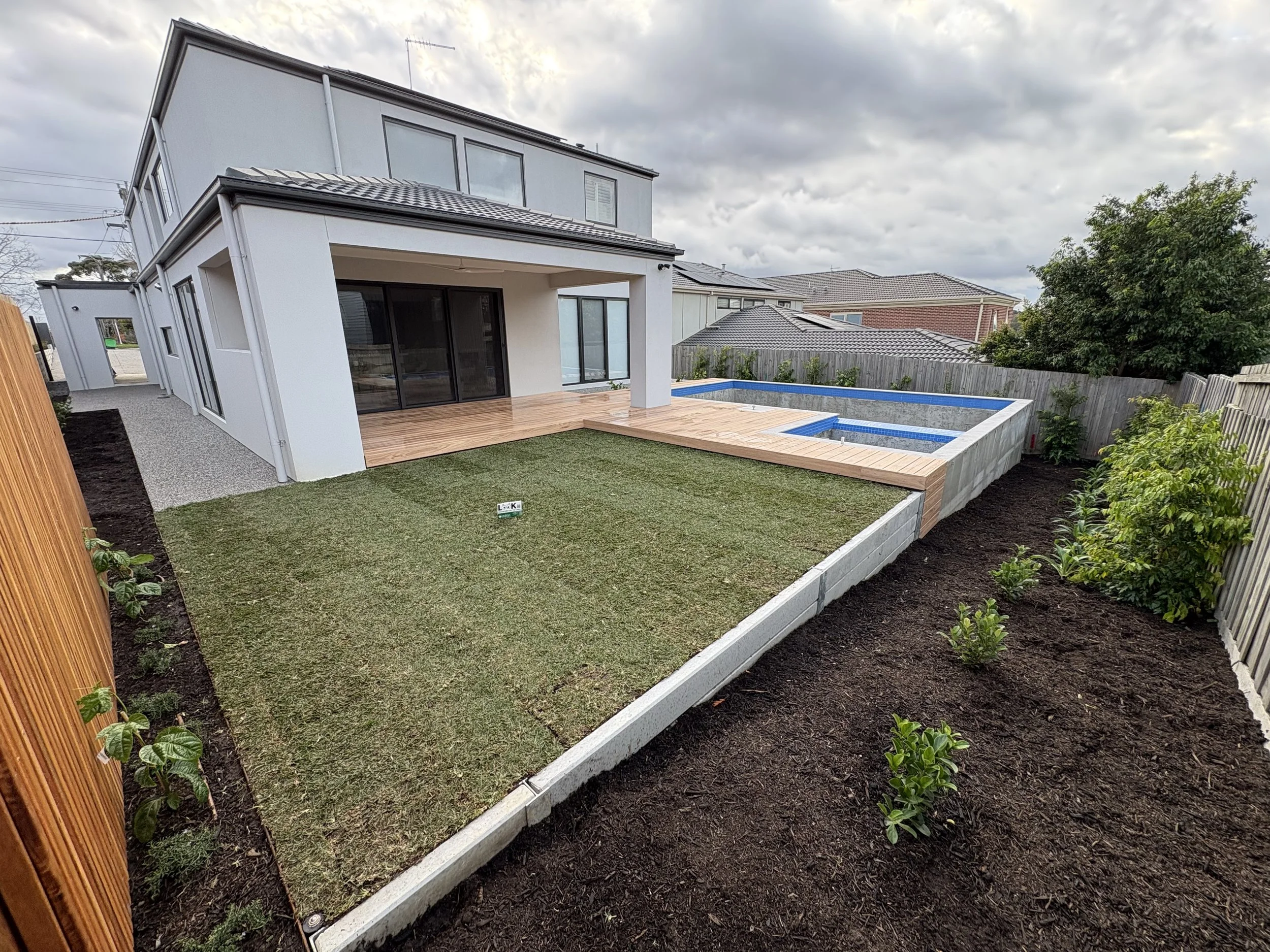 View of a modern two-story house with a backyard, featuring a wooden deck, an in-ground pool under construction, a grassy area, and surrounding shrubbery, with cloudy skies overhead.