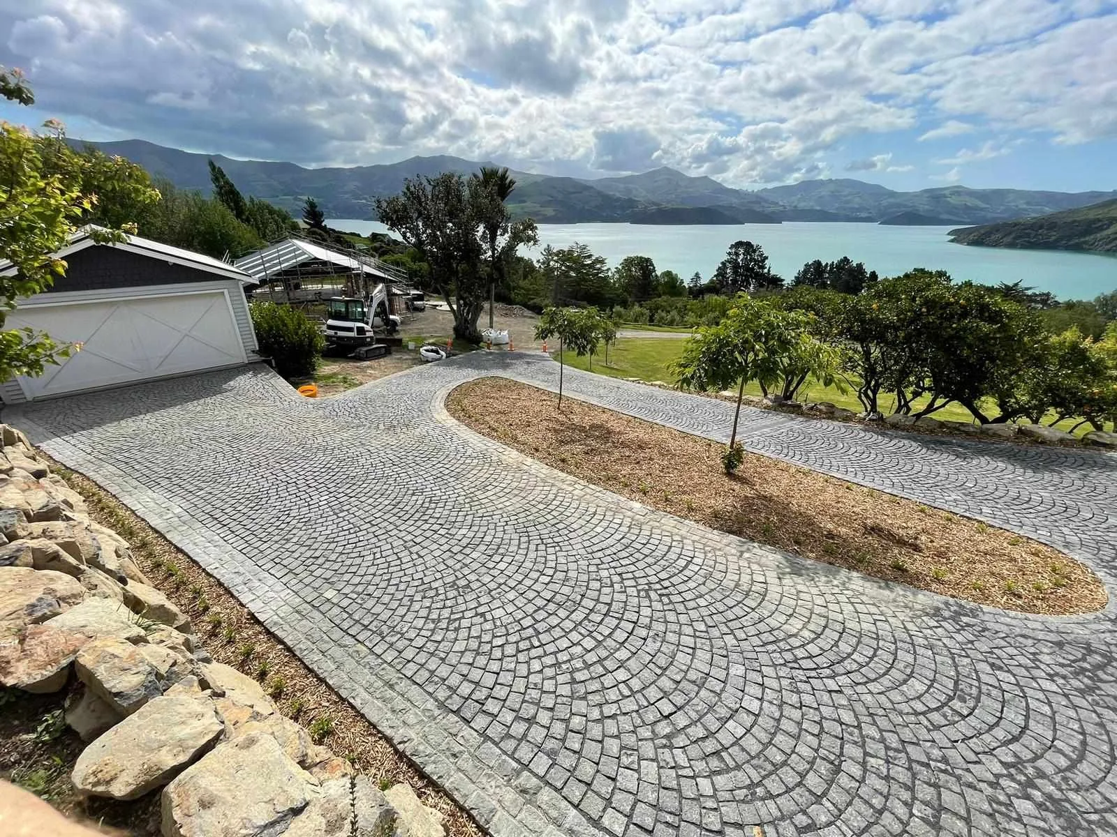 Landscape view of a paved driveway with a scenic lake and mountains in the background, trees, and partly cloudy sky.