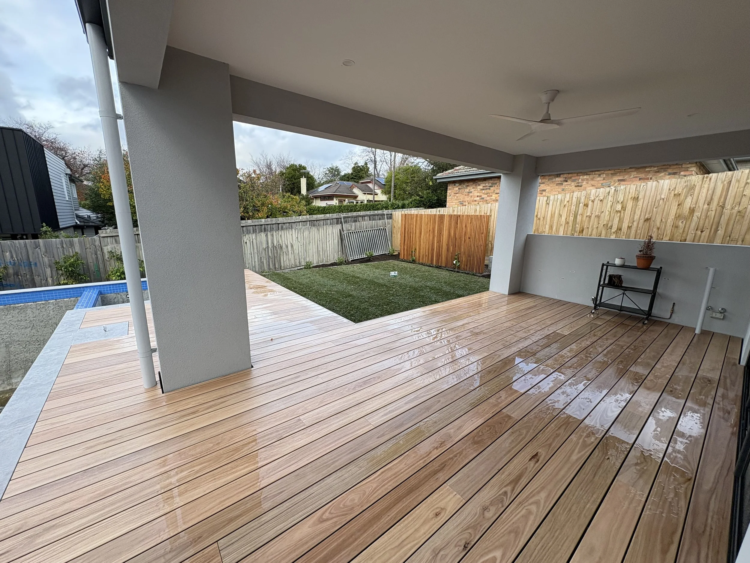 Covered outdoor patio with wet wooden floor, white ceiling fan, and a black metal shelf with potted plant, fencing, and neighboring rooftops visible beyond.