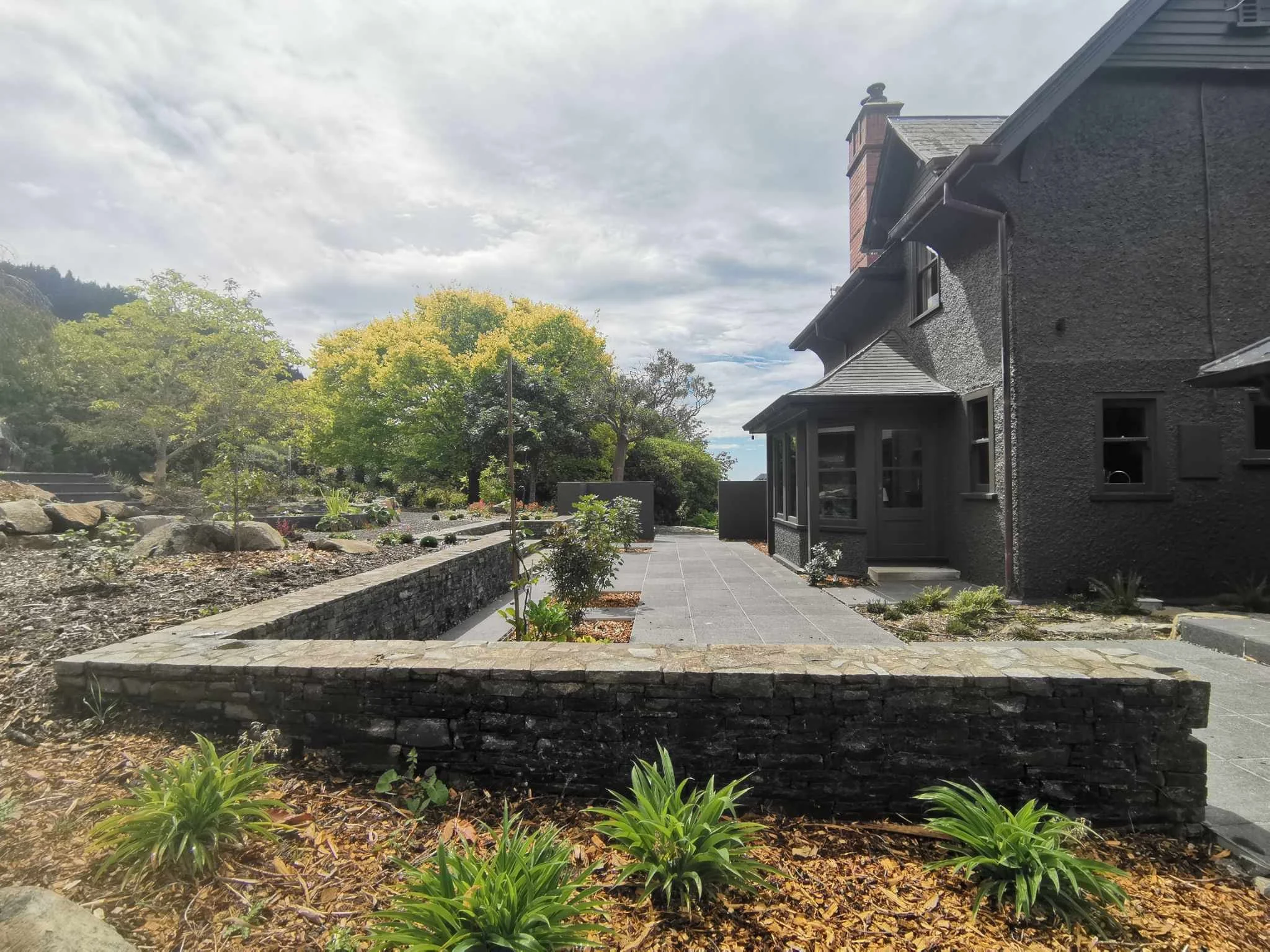 A backyard garden with a paved patio, green plants, and trees, adjacent to a gray house with a chimney, under a cloudy sky.
