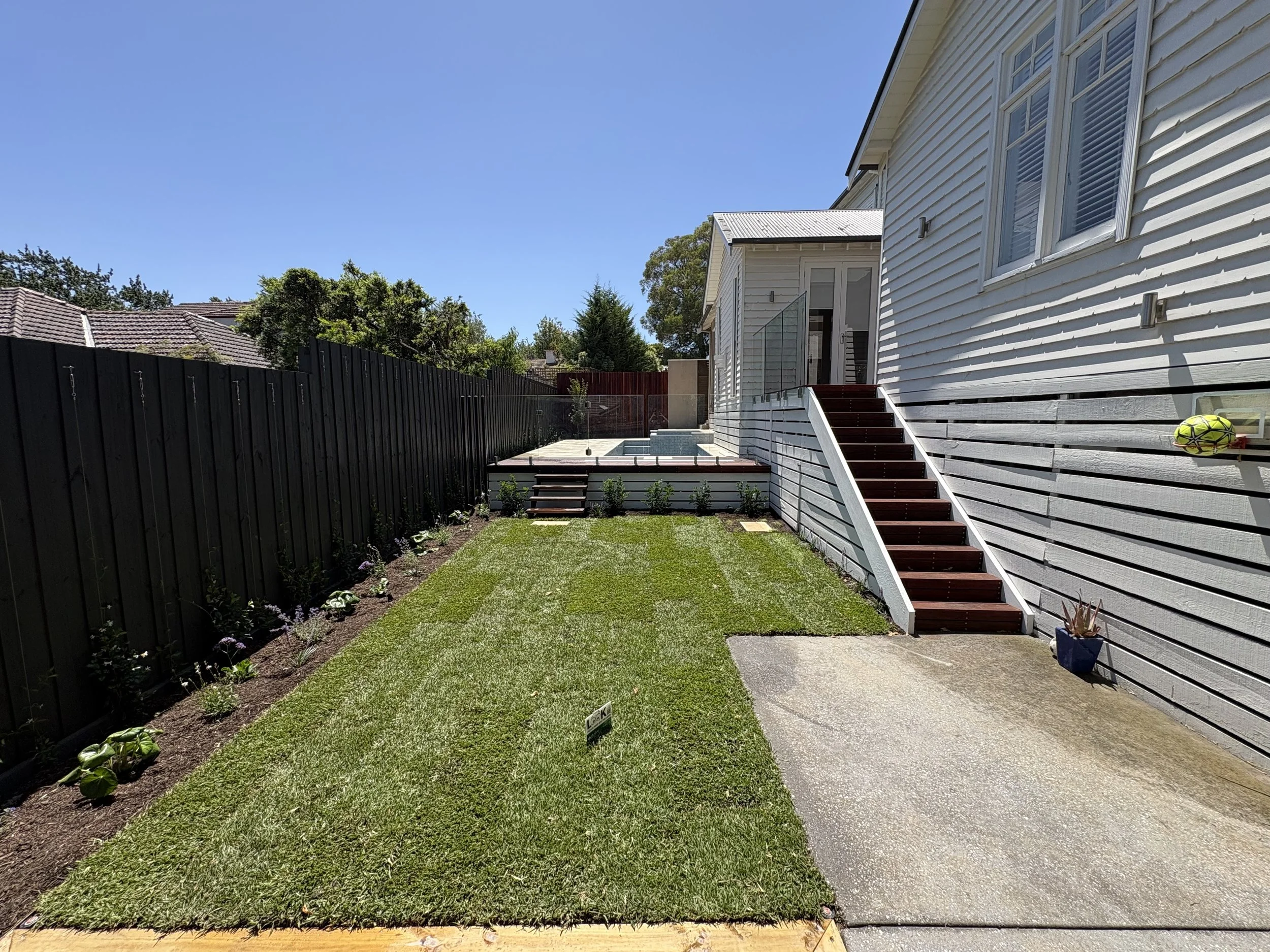 Backyard with green grass, flower bed on left, stairs leading to house deck, white house exterior with multiple windows, soccer ball mounted on wall, clear blue sky.