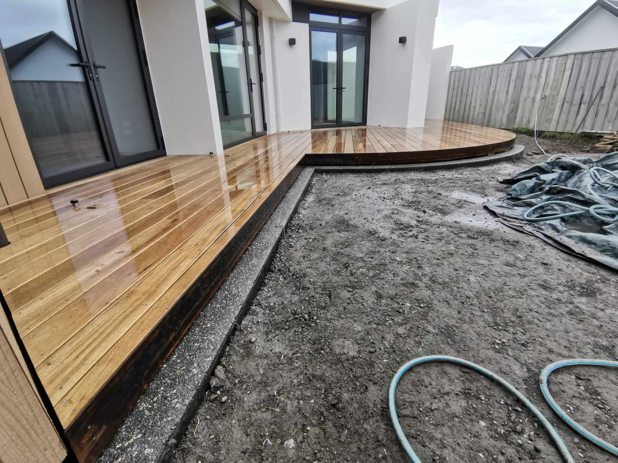A newly constructed house with a wooden deck outside, sliding glass doors leading into the house, and a gray cemented yard area with some pipes and construction materials scattered around.