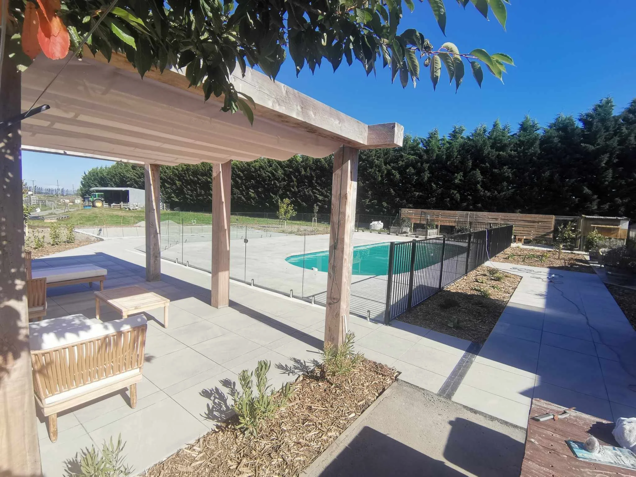 Backyard with a patio area, wooden pergola, outdoor furniture, swimming pool, and landscaped garden with trees and plants under a clear blue sky.