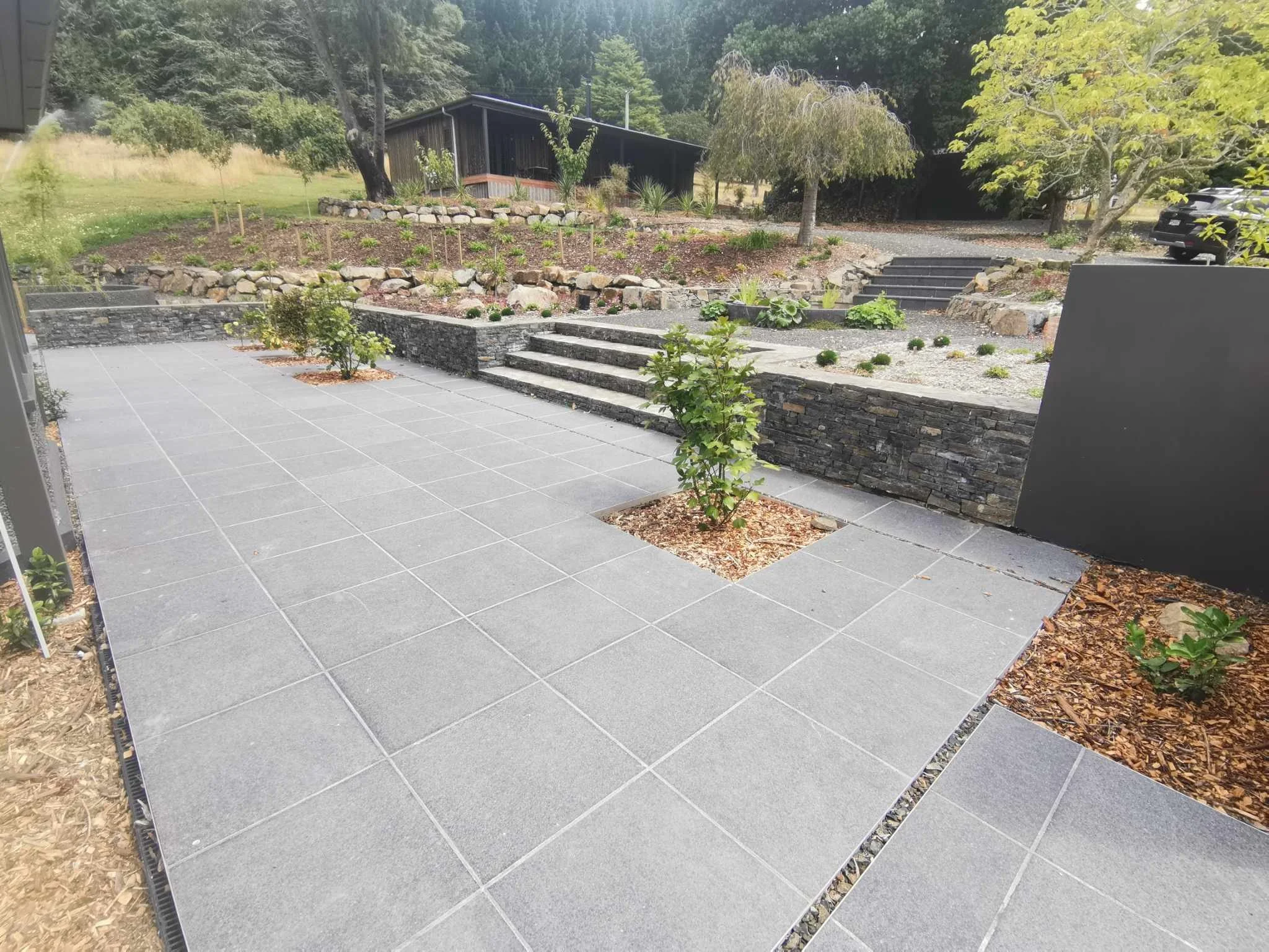 The image shows a modern backyard with a tiled patio, small trees planted in the ground, and a landscaped garden with steps leading to a higher level with plants and a dark wooden building in the background. There are trees and grass beyond the lands