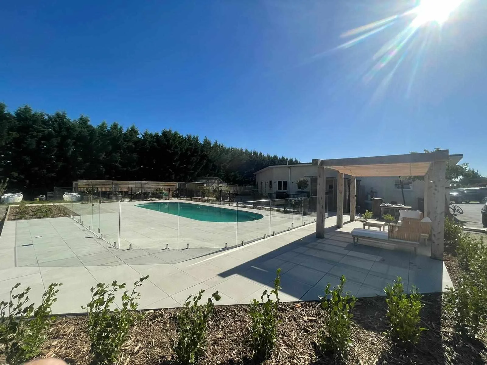 Residential backyard with a swimming pool surrounded by a glass fence, a shaded seating area with a wooden pergola, and a house in the background under a bright blue sky with the sun shining.