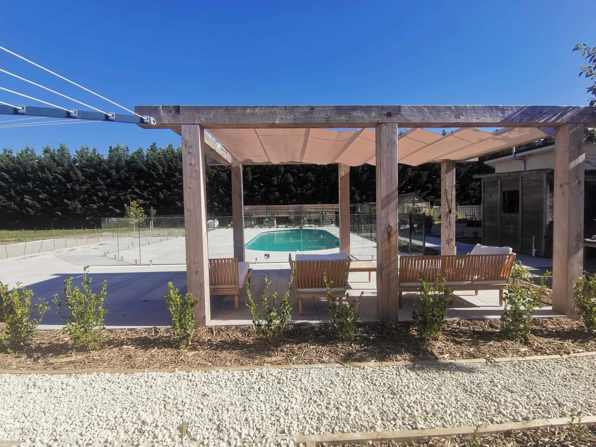A backyard with a wooden pergola, outdoor seating, a swimming pool, landscaping, and a clear blue sky.