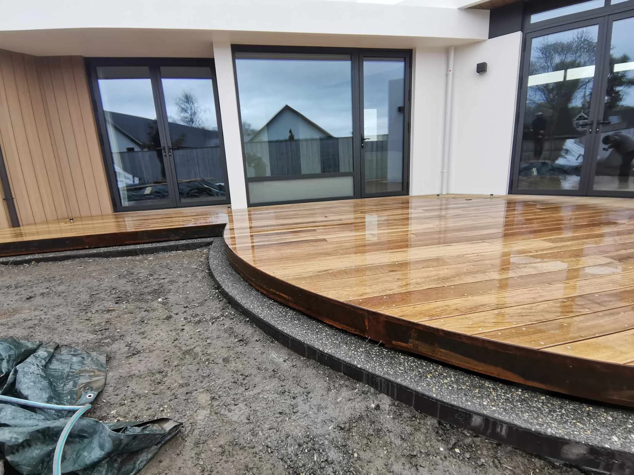 Newly built house with large glass sliding doors and a wooden deck, outdoor construction area with gravel and tools in front.