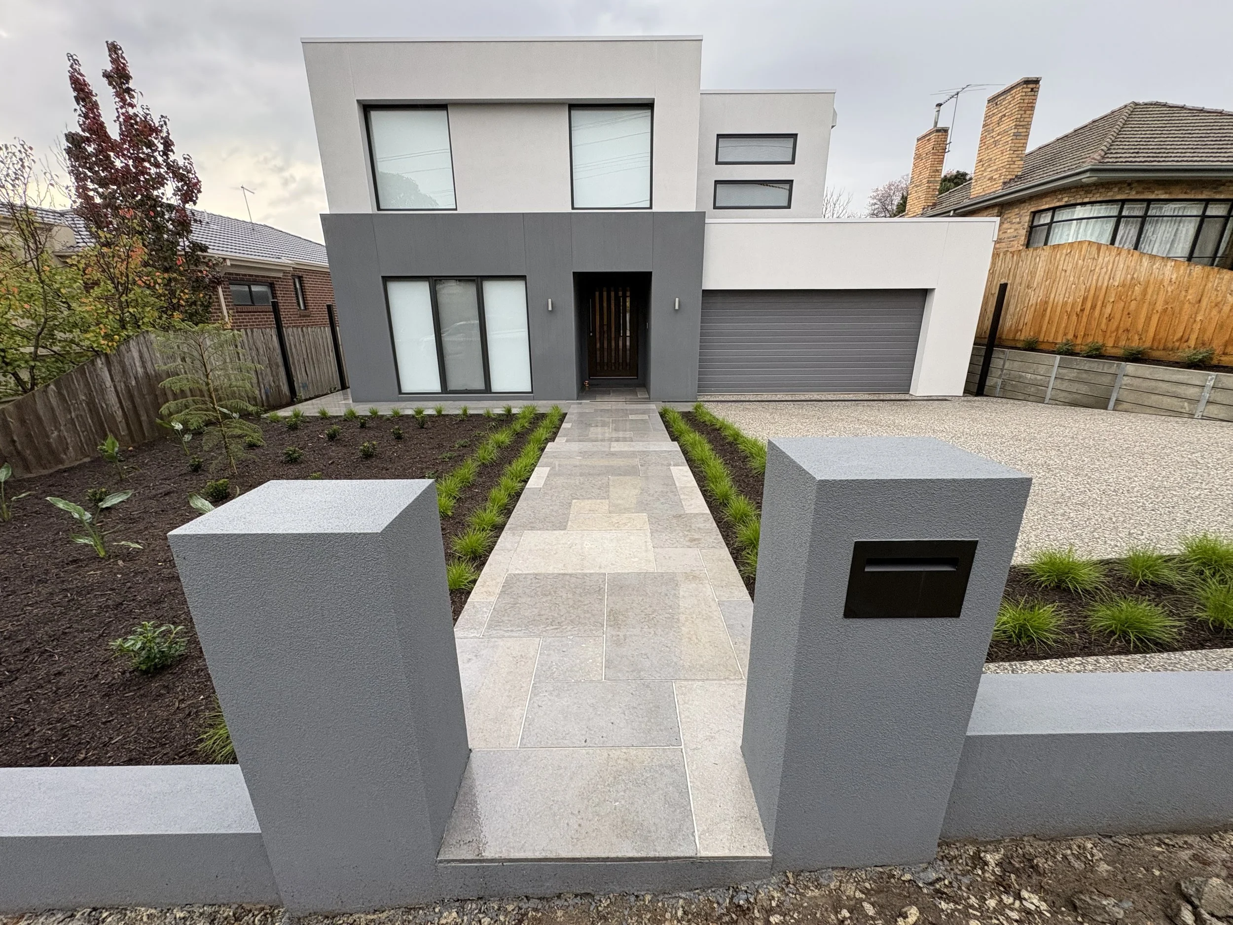 Modern two-story house with a white and gray exterior, large windows, a closed garage door, and a small landscaped front yard with young plants and a pathway.
