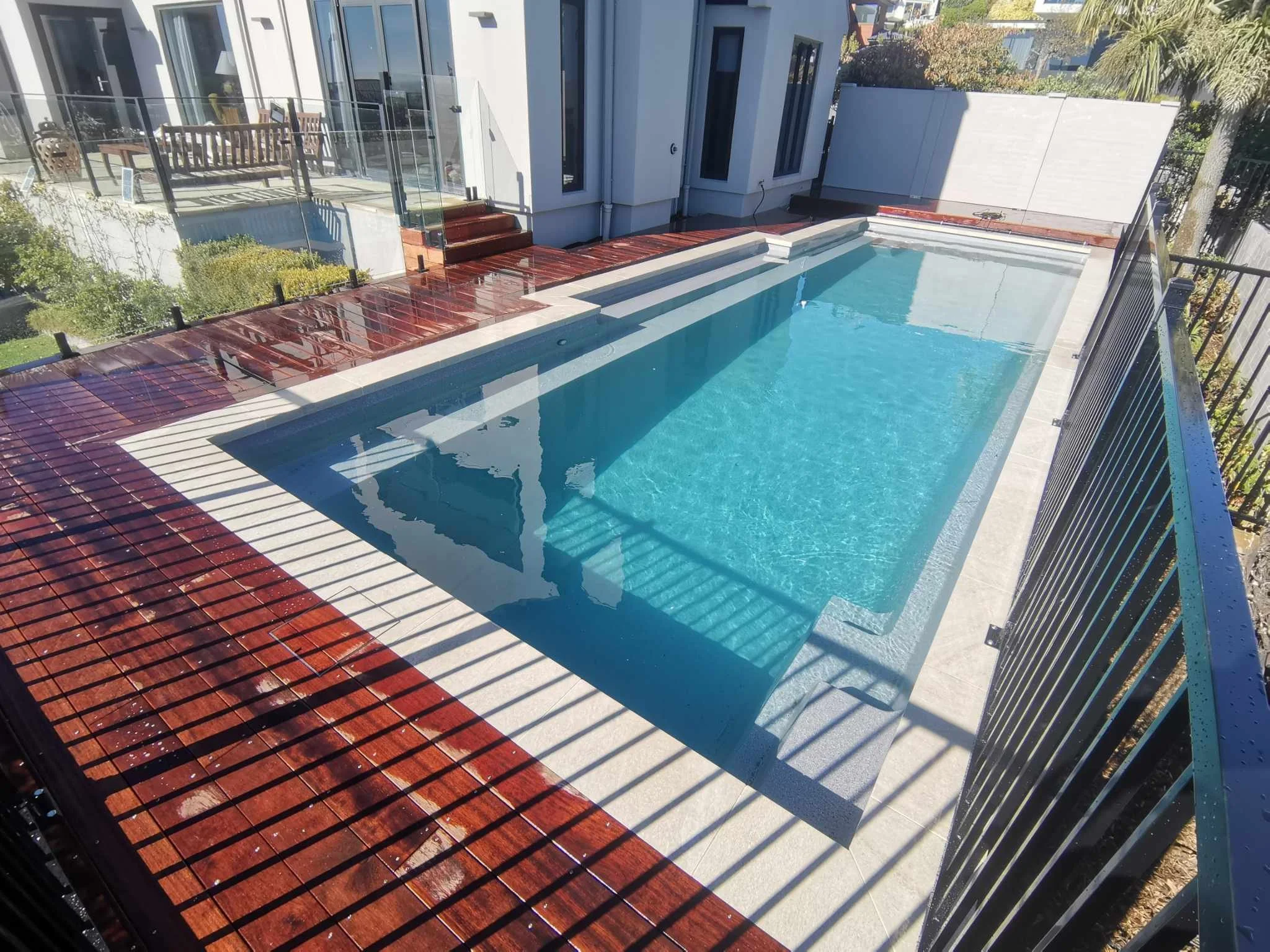 A backyard swimming pool with clear blue water, surrounded by a red wooden deck, with white steps leading into the pool, a white wall, and a garden with trees and bushes.