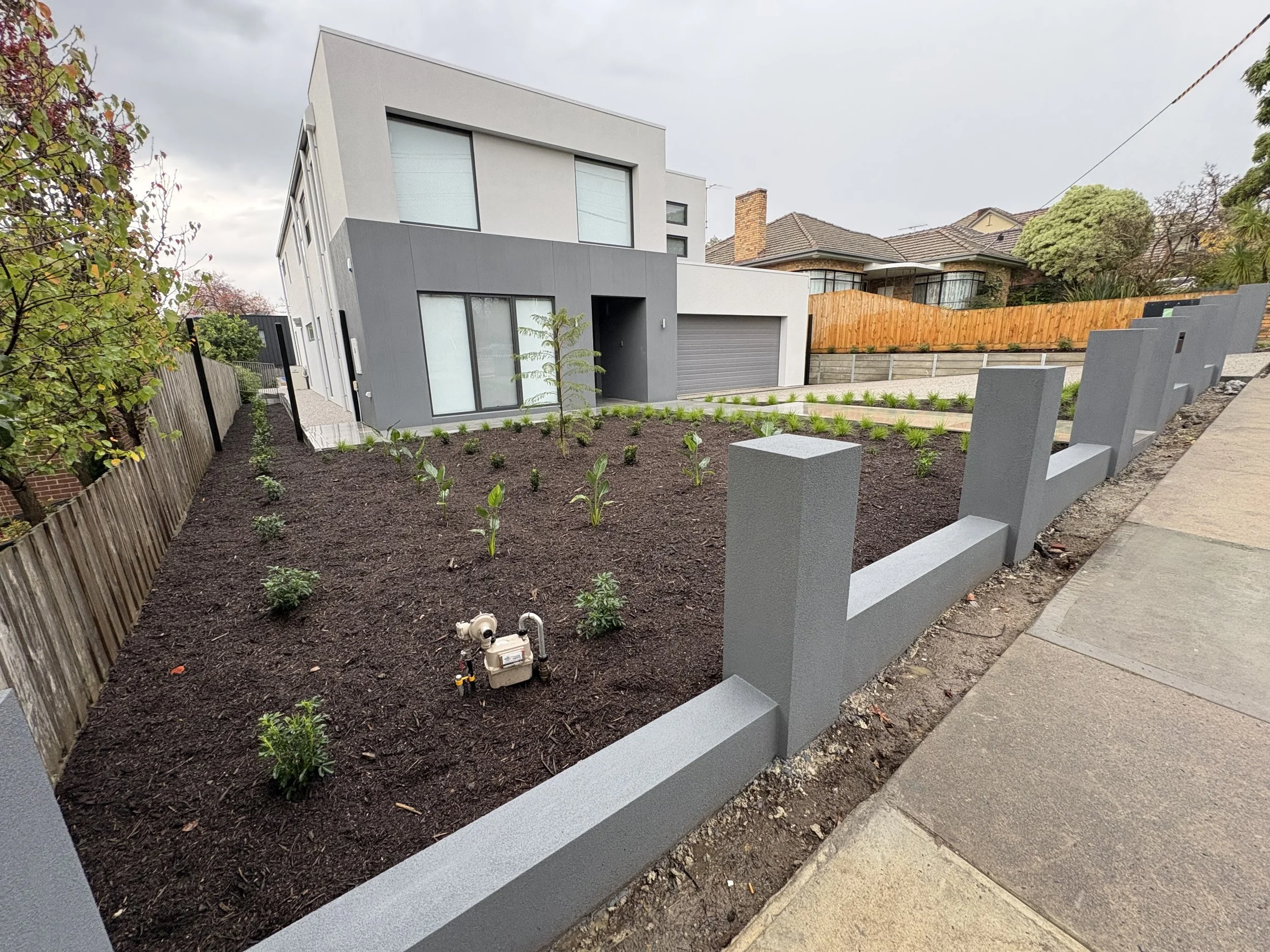 Modern house with a gray and white exterior, large windows, a sloped roof, and a small landscaped front yard with young plants, bordered by a concrete fence.