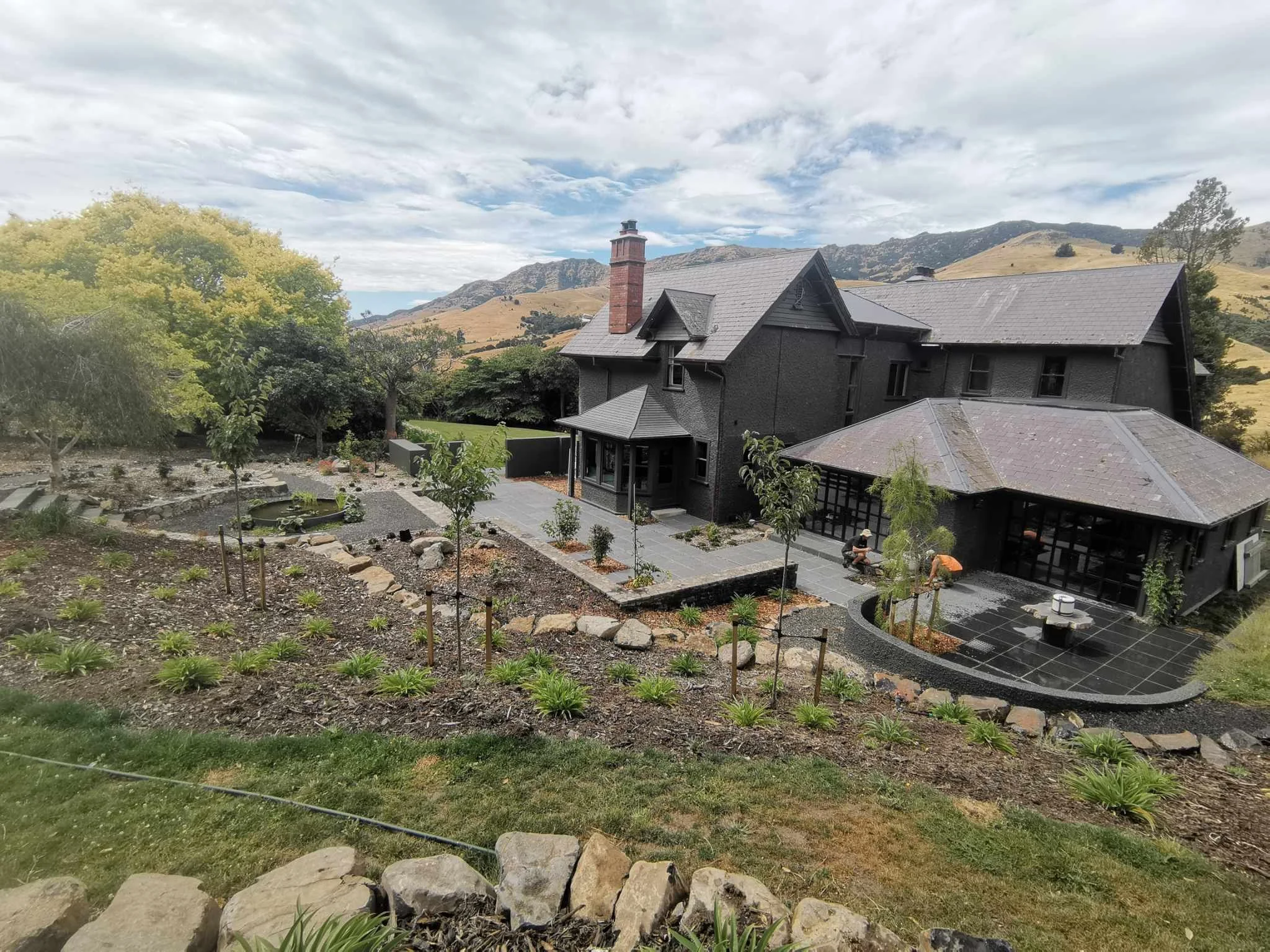 A house with dark exterior walls and gray slate roof, surrounded by a landscaped yard with young plants and trees, set against a backdrop of rolling hills and cloudy sky.