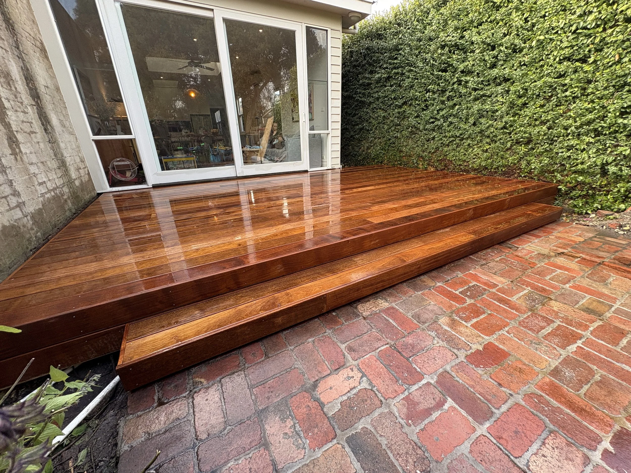 A polished wooden deck attached to a house with a sliding glass door, next to a brick patio and a green hedge.