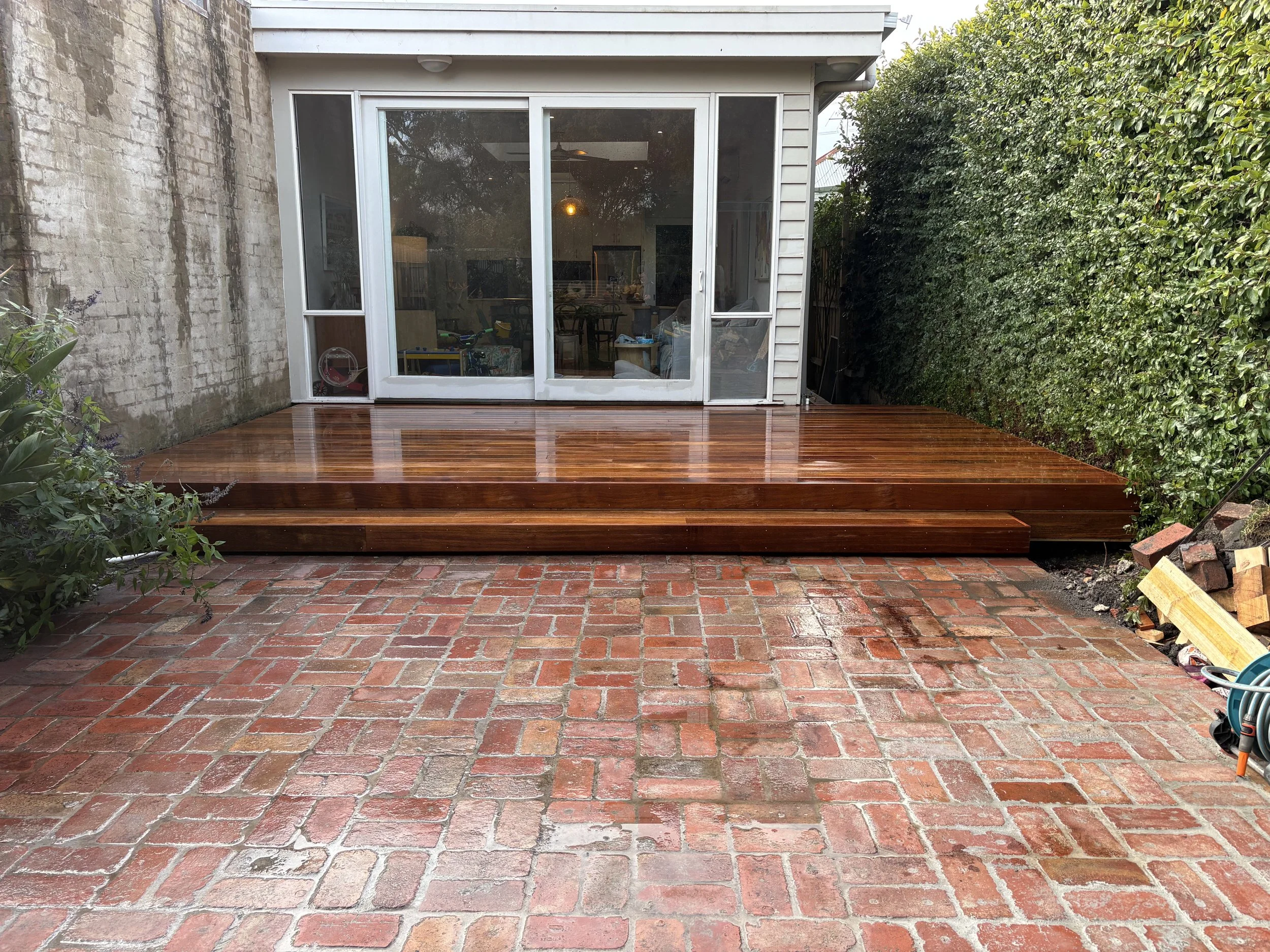 A backyard with a wooden deck, brick patio, a sliding glass door leading inside, a tall hedge on the right, an unfinished brick wall on the left, and gardening tools and materials on the ground.