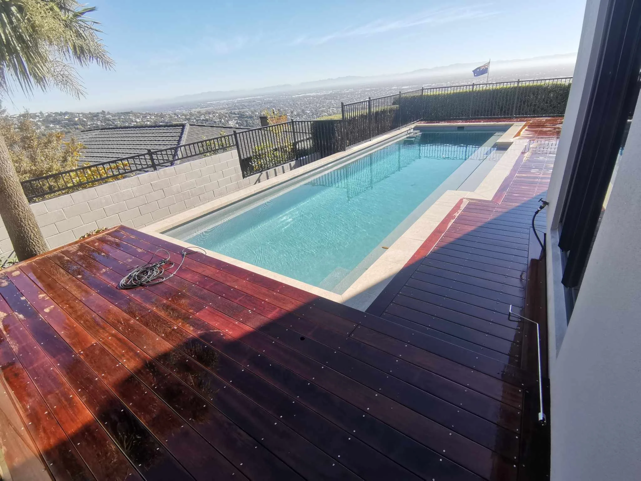A backyard with a rectangular swimming pool on a wooden deck, surrounded by a black metal fence, overlooking a cityscape and a flagpole with an Australian flag in the distance.