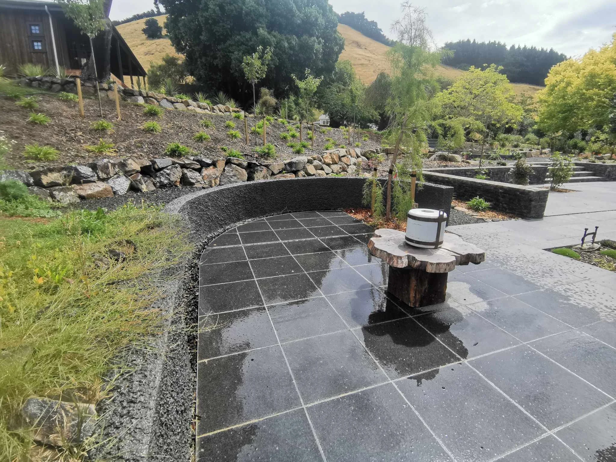 A landscaped outdoor area with a patio, decorative stone edging, small plants and trees, and a background of hills and trees.