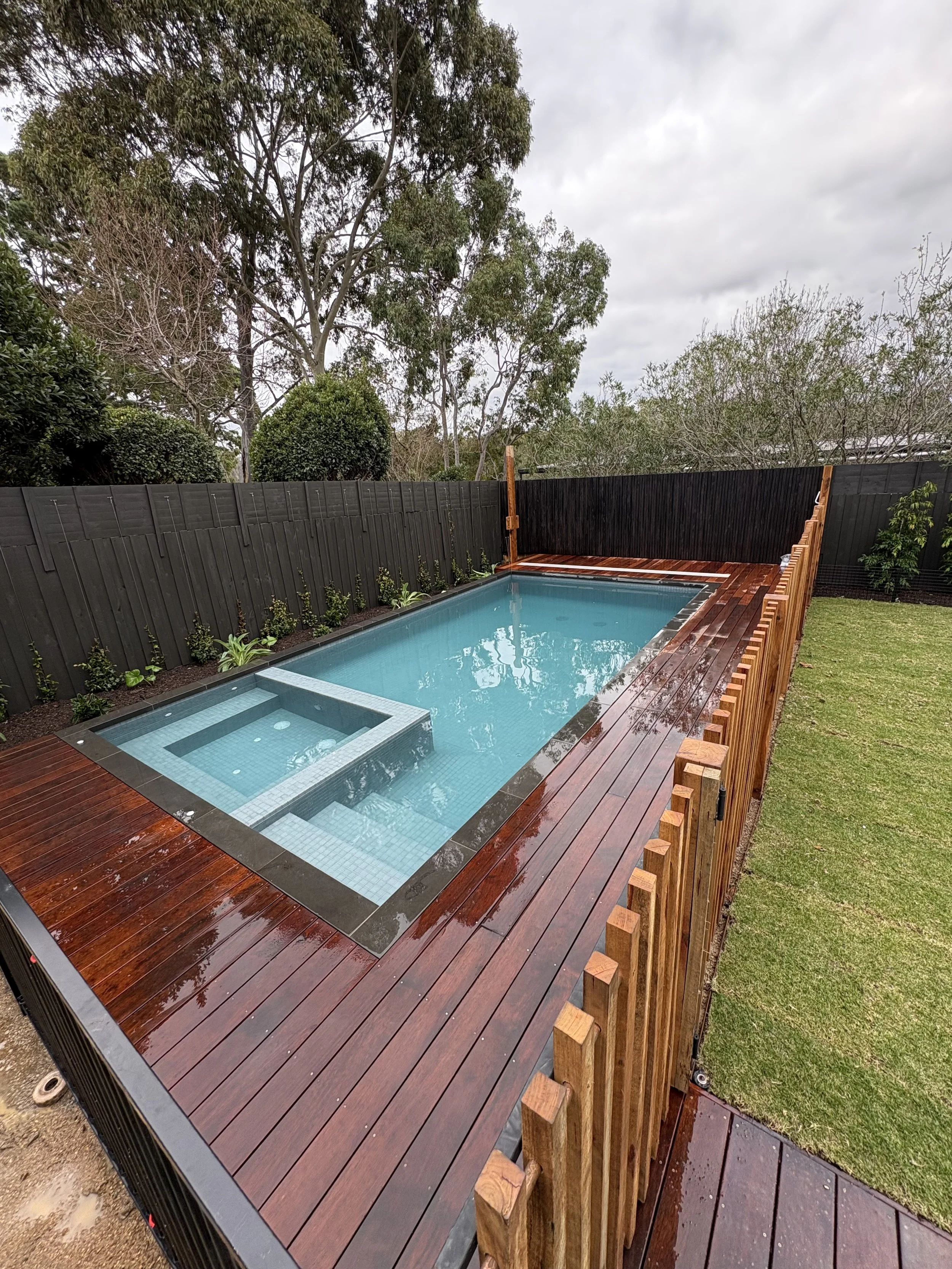 A backyard view featuring a rectangular swimming pool with clear water, surrounded by a wooden deck and a black privacy fence. In the background, there are tall trees and cloudy sky, with some greenery along the fence line.