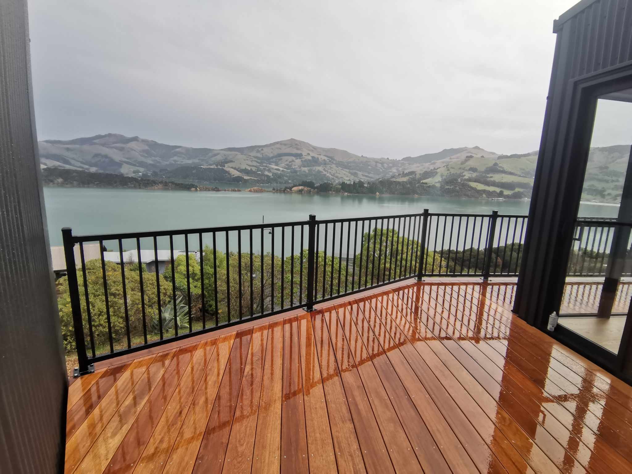 A wet wooden deck with orange-brown planks and black metal railing, overlooking a body of water with green hills and mountains in the background under a cloudy sky.