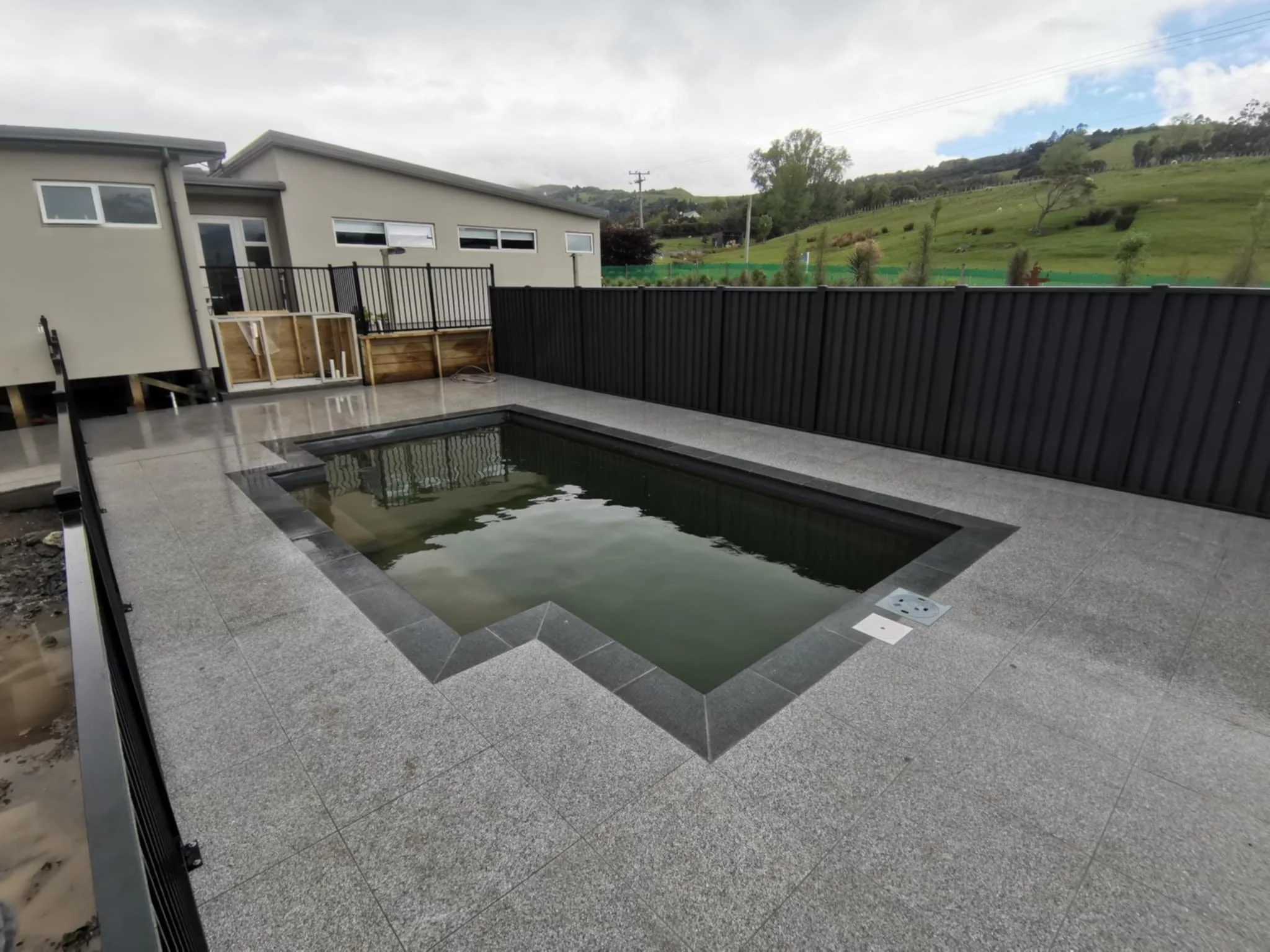 A backyard with a small pool, gray stone patio, black fence, and a background of green hills and trees under a cloudy sky.
