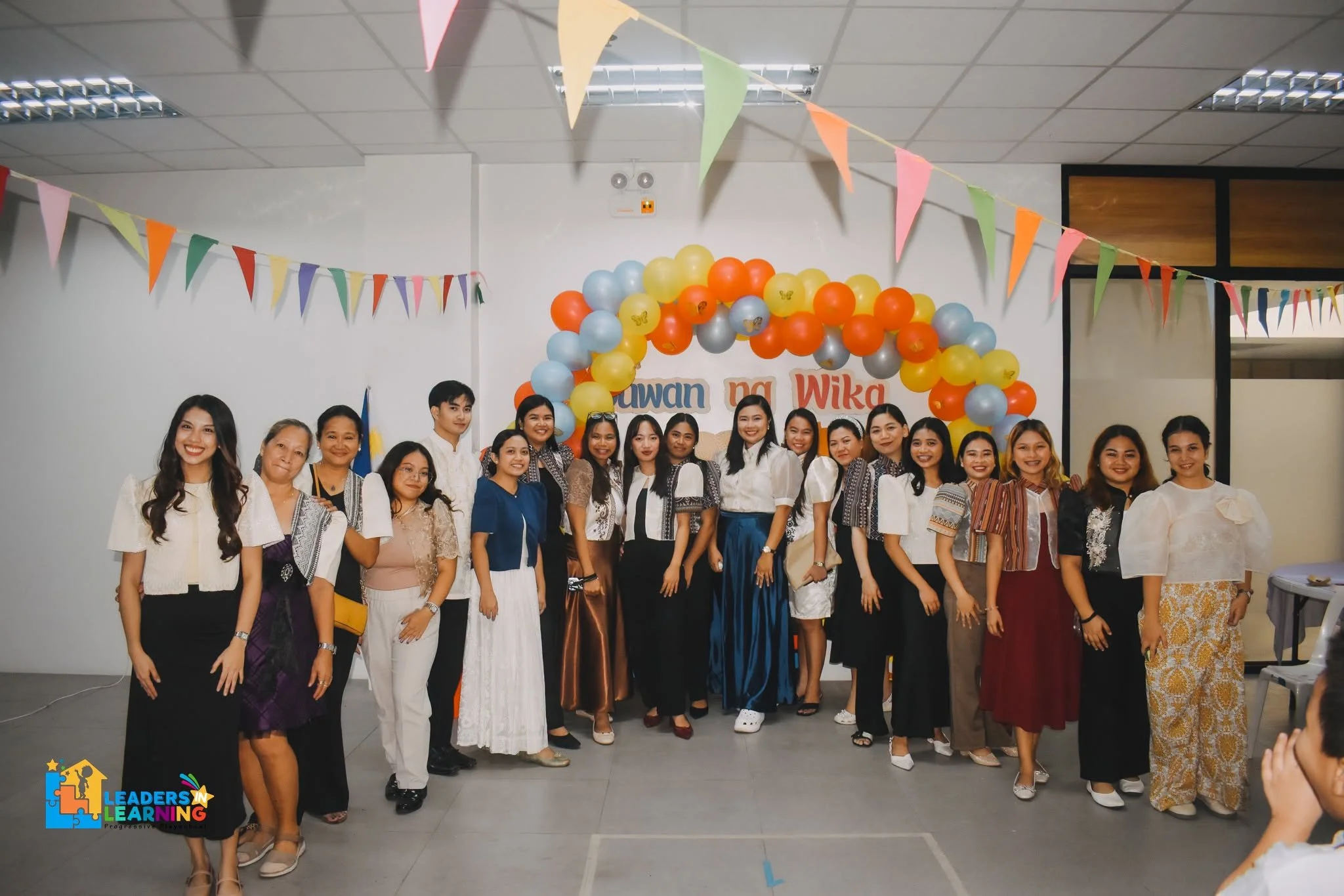 A group of diverse women posing together in front of colorful balloon arch and banners at an indoor event. The women are smiling and dressed in casual and semi-formal attire. The background has decorations with the words 'Dawan na Wika' and a logo in the bottom left corner indicating a learning program.