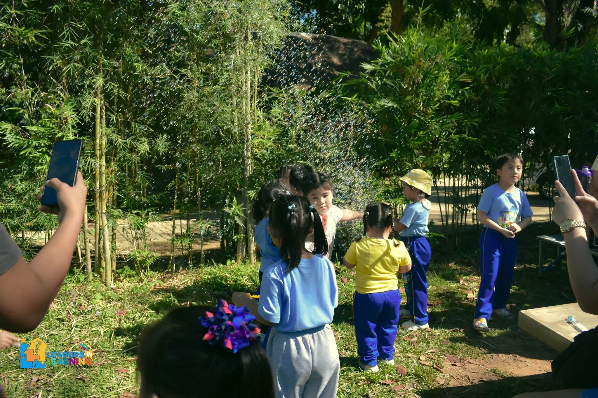 Children gathered outdoors near green bushes, with some taking photos or videos, and a lady in a yellow hat in the background.