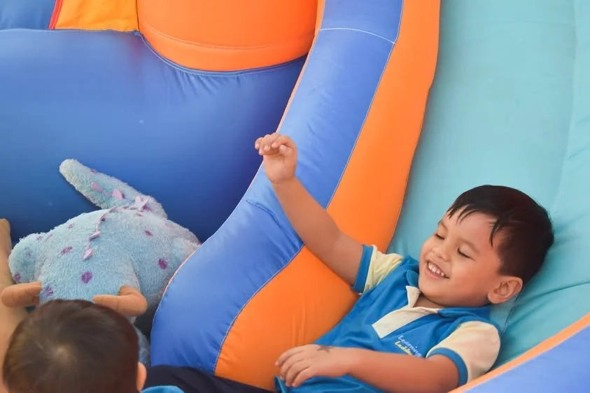 A young boy with dark hair, smiling and reaching up inside an inflatable play area, with a plush unicorn toy nearby.